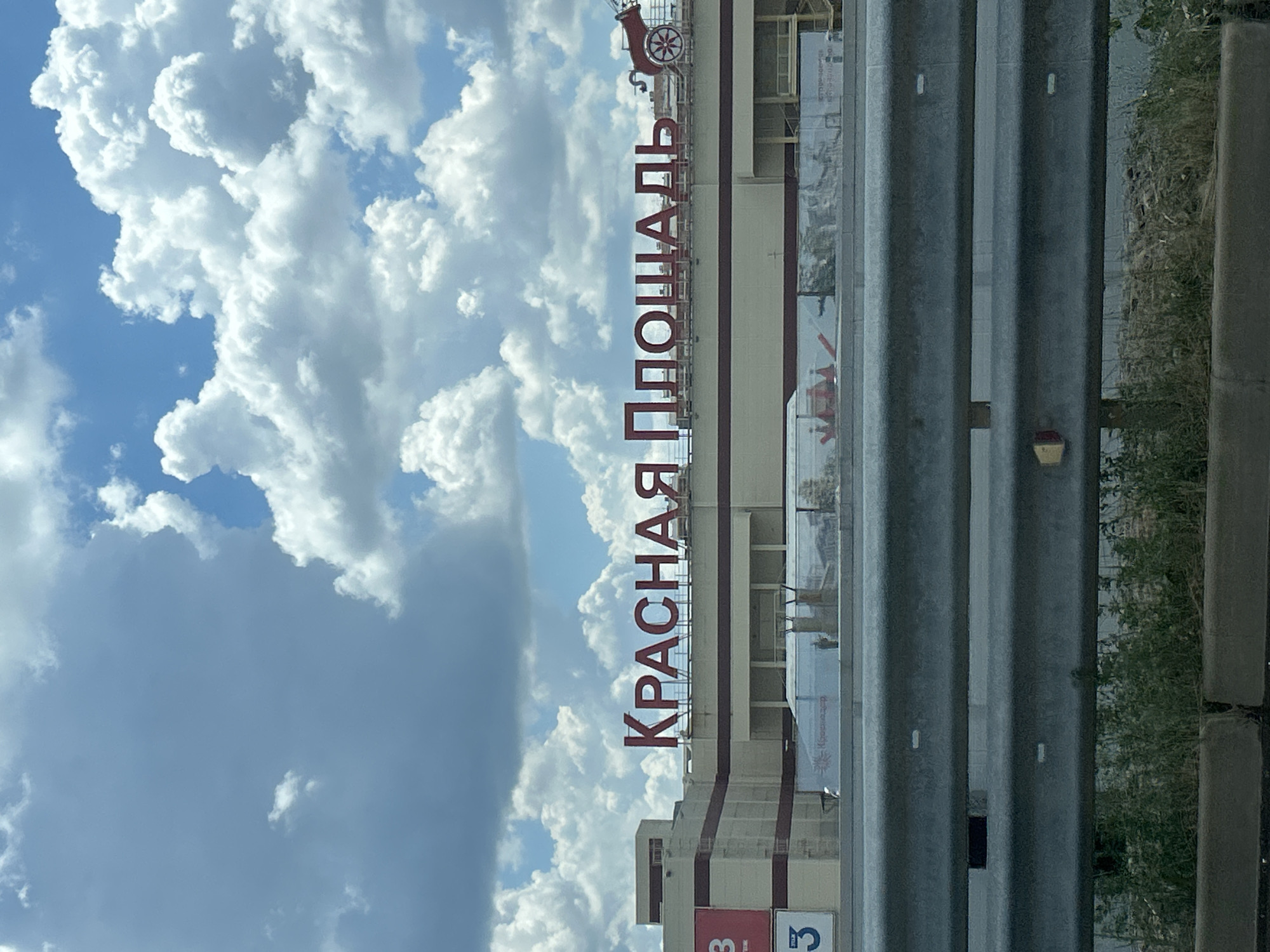 The image shows a building with a sign that reads "КРАСНАЯ ПЛОЩАДЬ" (Red Square) in large red letters. The building appears to be a commercial establishment. In the foreground, there is a metal guardrail, and behind it, some vegetation and concrete barriers. The sky is visible above the building, with clouds of varying densities. There are no people or animals clearly visible in the image. The context of the building and its sign suggests it might be a shopping center or a similar public space. The city of Orlovskii, Russia is mentioned as the location.