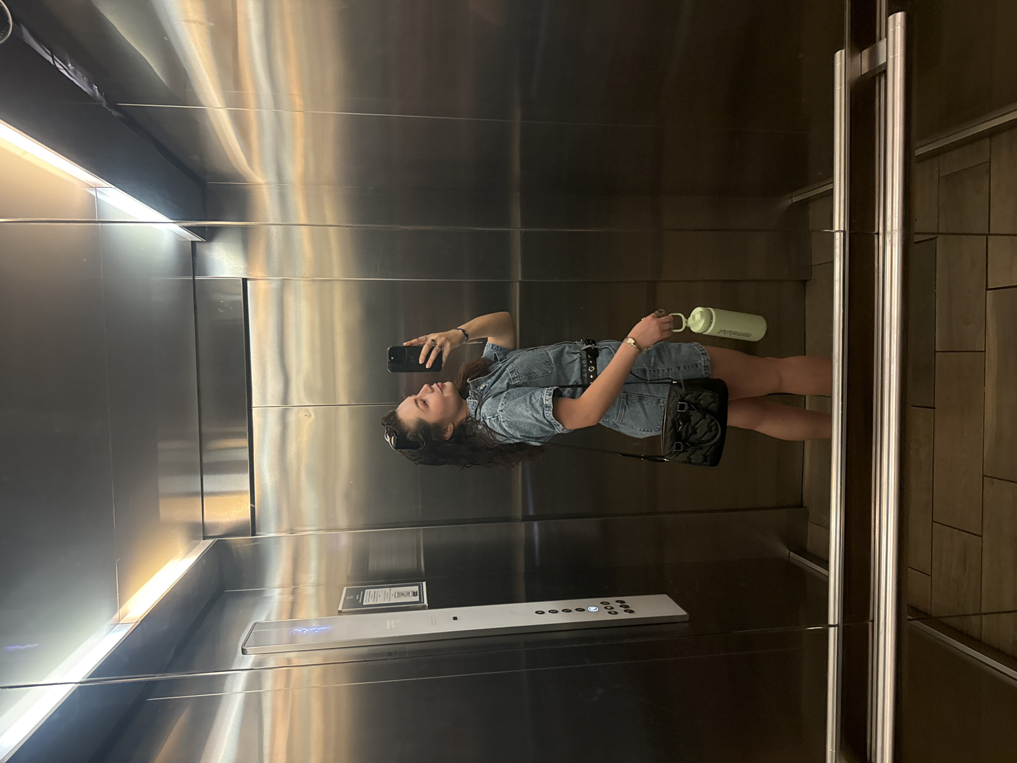 An overhead-angle, indoor shot depicts a young woman in a reflective elevator, who is presumed to be taking a selfie with her cell phone. She is holding a green water bottle in one hand and the cell phone in the other. The woman wears a denim dress with a belt, has curly dark hair, and is wearing sunglasses atop her head. A small black purse hangs from her shoulder.
The interior of the elevator has a polished metal finish, creating reflections of the lighting and the elevator's controls. The elevator buttons and a small informational sign are visible near the bottom of the shot. The elevator door frame and a tiled wall are also present on the right side of the image. The lighting is bright and possibly fluorescent.
No information suggests that the location is in Mexico City, Mexico.