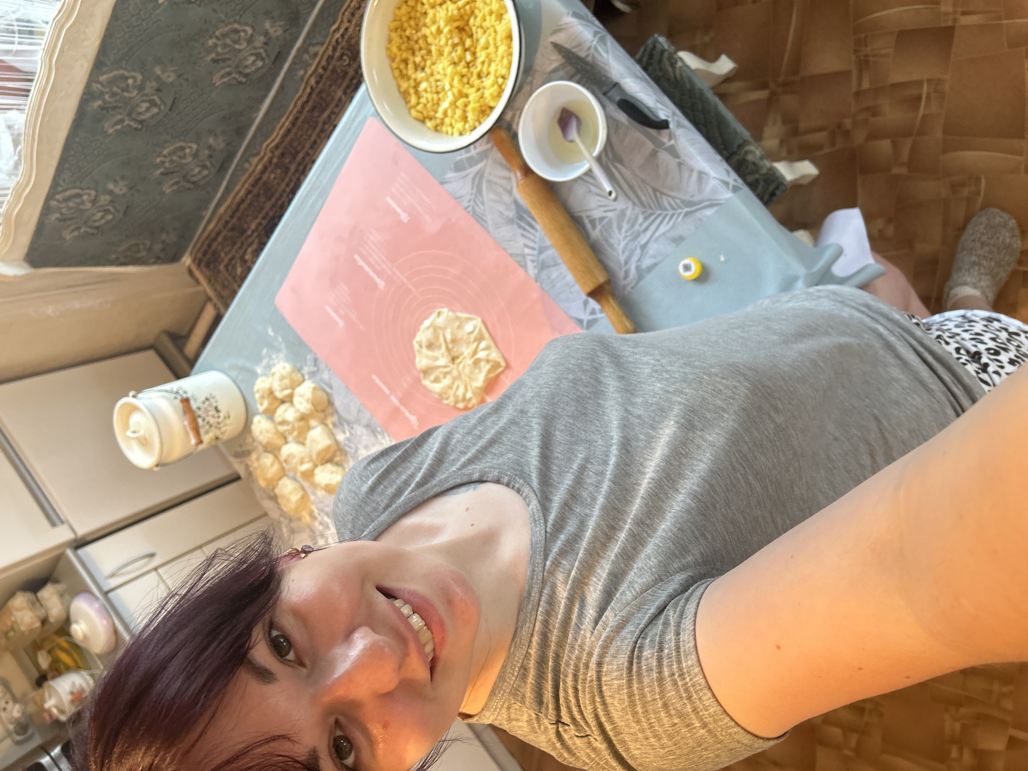 This image, taken in a kitchen in Фоки, Russia, depicts a woman engaged in food preparation.
The subject, a woman with dark reddish-brown hair and light skin, smiles directly at the camera. She wears a grey short-sleeved t-shirt and patterned shorts, with grey textured slippers visible on her right foot. An earring with small purple beads hangs from her left ear. Braces are visible on her upper teeth.
The scene is dominated by a light blue table with a white and grey leaf-patterned tablecloth. On the table, various items related to cooking are arranged:
- A pink silicone mat, marked with measurements and text in what appears to be Cyrillic, displays a partially formed piece of dough, pleated into a flower-like shape.
- To the left of the mat, several small, rounded pieces of dough are arranged on a light surface, possibly parchment paper or a floured cloth.
- A white bowl filled with diced yellow ingredients, likely fruit or apples for a filling, is positioned on the right side of the table.
- A wooden rolling pin, a small white bowl containing a clear liquid (possibly egg wash or oil) with a small spoon, and a knife with a black handle are also present on the tablecloth.
- A small yellow cap or lid is near the bottom center of the frame.
In the background, a kitchen counter and light-colored cabinets are visible. A large, white cylindrical container with a floral pattern and a lid sits on the counter. Further back, a window with a patterned, possibly blue and grey, fabric valance or curtain can be seen. A dark patterned rug is visible on the floor beneath the window. The floor is tiled in a light brown with a geometric pattern.