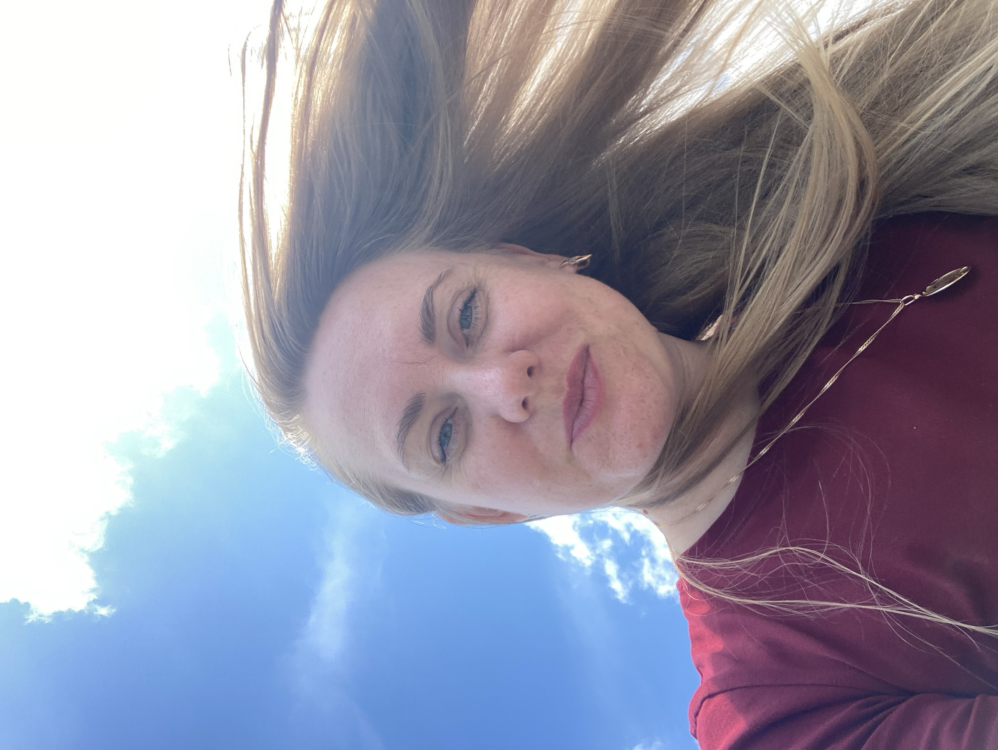A woman with blonde hair is taking a selfie outdoors. She is wearing a maroon shirt and a necklace. Her hair is blowing in the wind. The background is a bright blue sky with some clouds. The image was taken in Марковский, Russia.