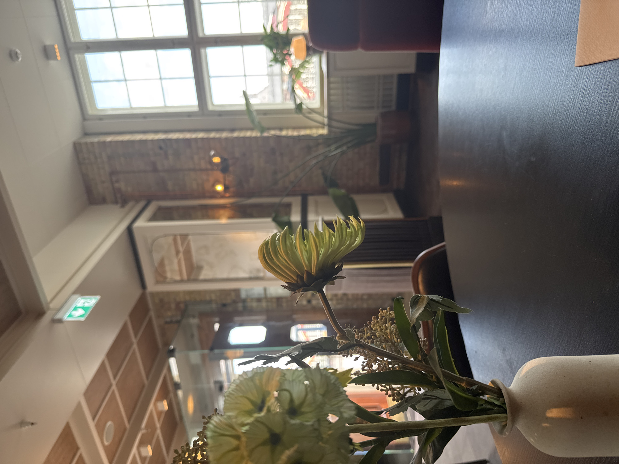 An indoor shot captures a close-up of a light-colored vase containing a floral arrangement of light green and pale yellow flowers and greenery. The vase sits on a dark-surfaced table. In the background, through large windows with white mullions, a brick building and outdoor scenery with red and orange umbrellas are visible. An indoor plant in a brown pot stands near the window. The room features exposed brick walls, a wooden-paneled ceiling with inset lighting, and a green illuminated exit sign. The overall impression is that of a restaurant or cafe setting.