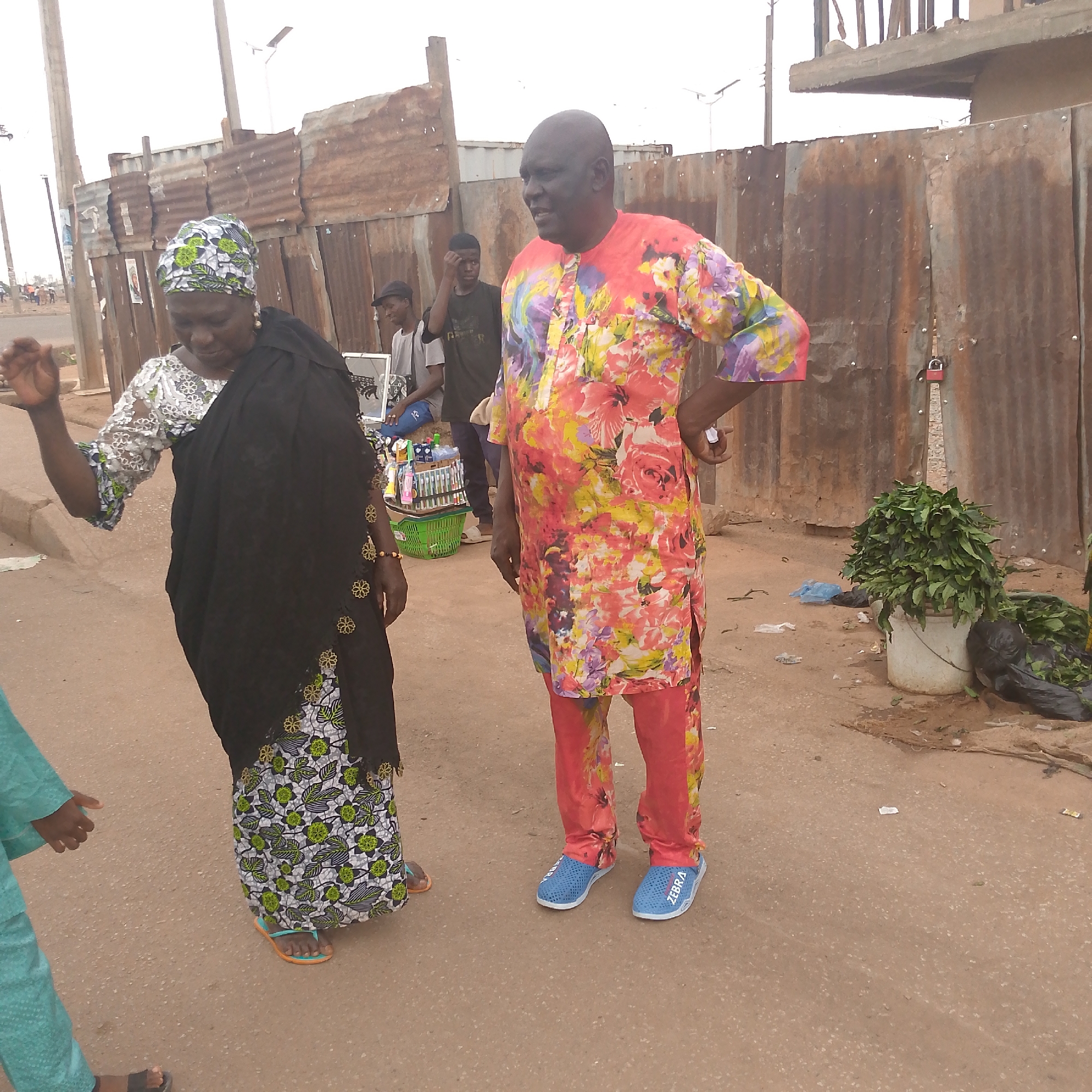 The image shows two individuals standing on a paved surface in Bukuru, Nigeria, with a corrugated metal fence behind them. The woman on the left wears a patterned dress, a black shawl, and a head covering. She is gesturing with her right hand. The man on the right is dressed in a brightly colored floral-patterned outfit with matching pants and blue shoes. He has his left hand on his hip. In the background, there are other people, a vendor's cart displaying various items, and a bucket of green leaves. The location appears to be a street scene.