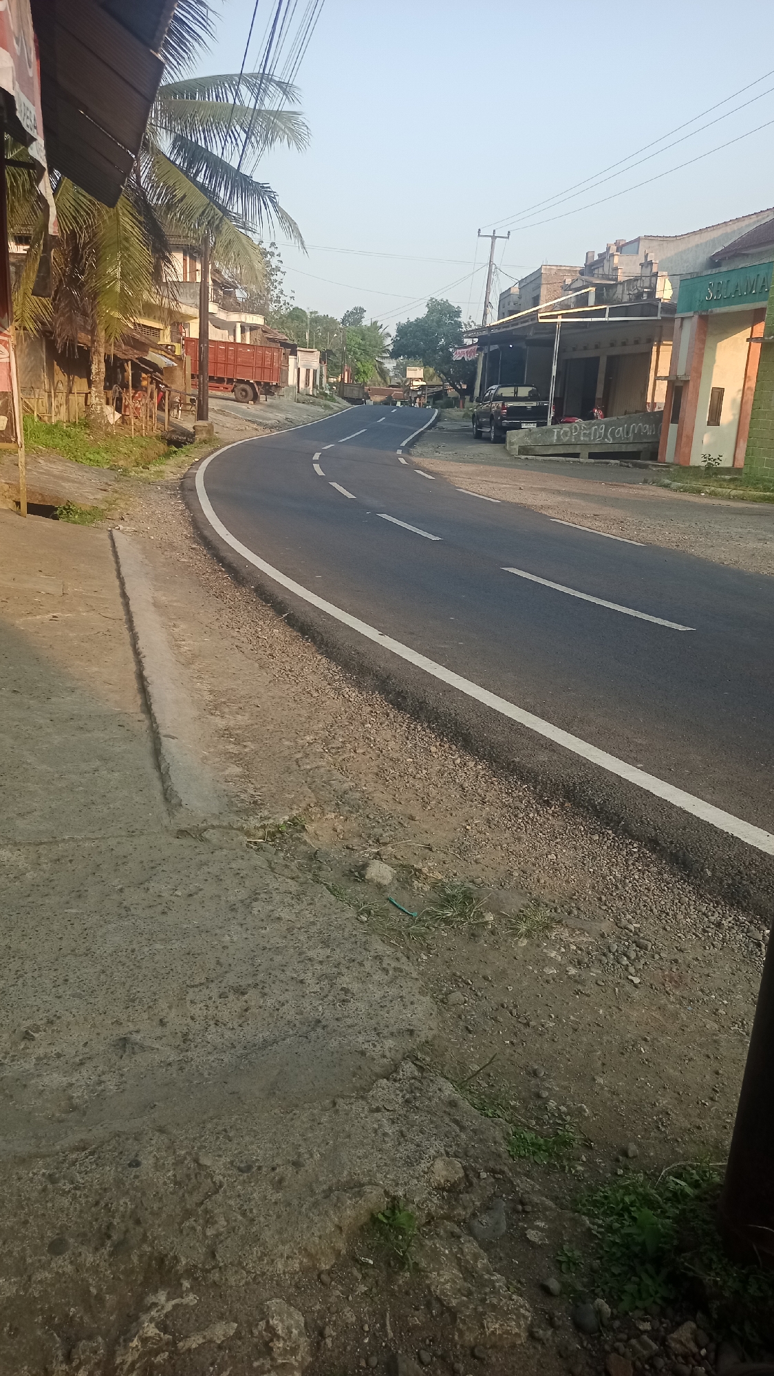 The image depicts a street scene in Cogreg, Indonesia, under clear skies and soft daylight. A paved road curves through the scene, bordered by a gravel shoulder on the right and a concrete sidewalk on the left. Various structures line the road, including low-rise buildings with awnings, and a prominent red truck is parked on the left side. A few parked vehicles are visible on the right. The overall impression is that of a typical roadside environment in a rural or semi-urban Indonesian setting, possibly in the morning or late afternoon given the lighting.