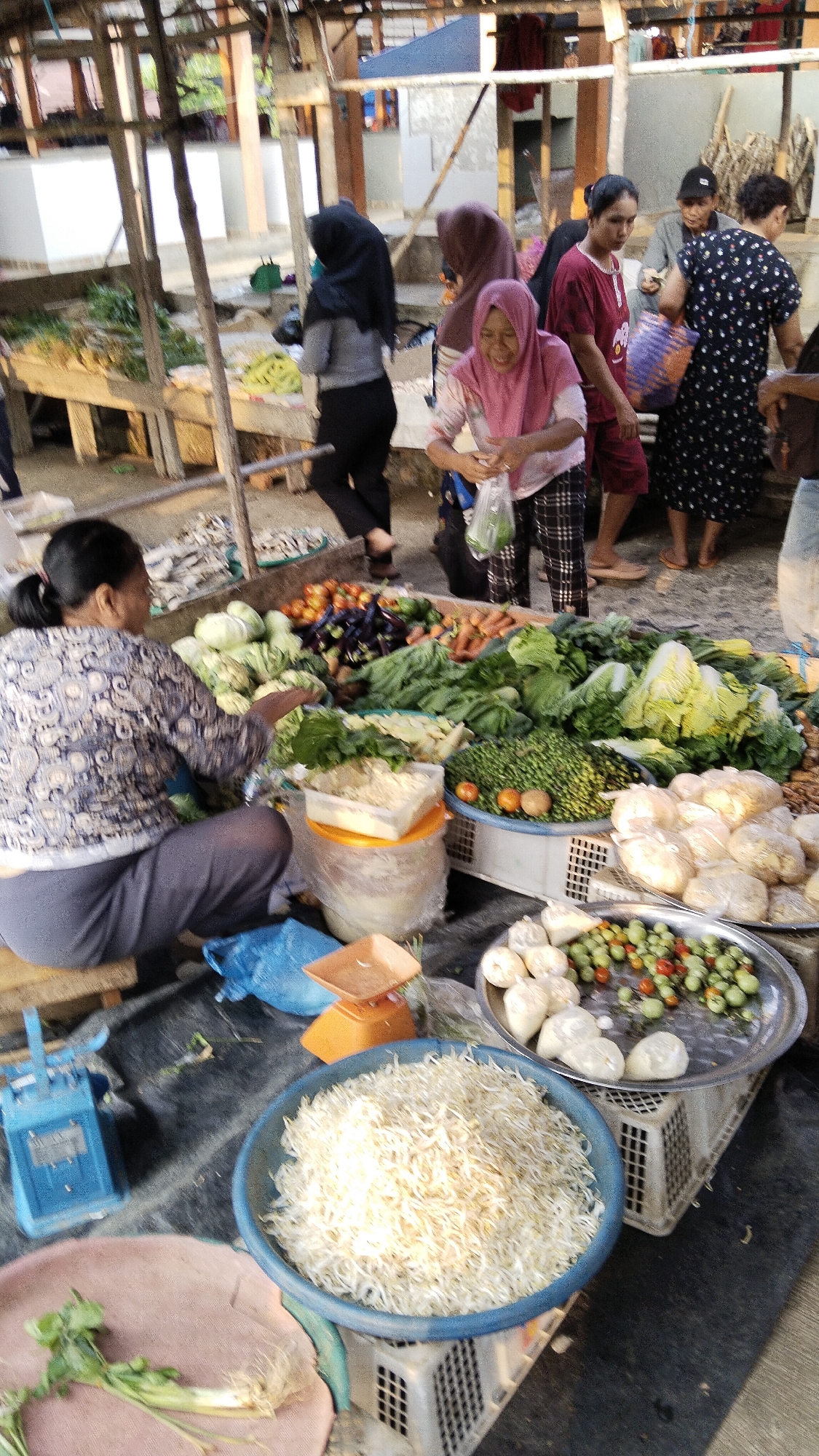 This image depicts a bustling outdoor market scene, likely in Margatunggal, Indonesia, with a variety of fresh produce and several individuals engaged in commerce. In the foreground, a woman wearing a patterned blue and white shirt and grey pants is seated on a wooden pallet, tending to a display of vegetables. A blue scale is visible to her left, and a blue plastic bag and a small orange scale are nearby. In front of her, a large blue basin is filled with a mound of what appears to be dried anchovies or similar small fish. Adjacent to this basin, a round metal platter holds small green and red cherry tomatoes along with some rounded white items, possibly garlic or onions, all resting on a white plastic crate. Further back, a vibrant assortment of vegetables including cabbage, leafy greens, tomatoes, and carrots are displayed on a stall. Several women, dressed in modest attire with headscarves, are interacting around the stall, some holding plastic bags and appearing to be customers or vendors. The market is set up under a wooden canopy, suggesting an open-air environment typical of Indonesian markets. The ground is covered with a dark tarp or mat. The overall atmosphere is one of lively trade and community engagement.