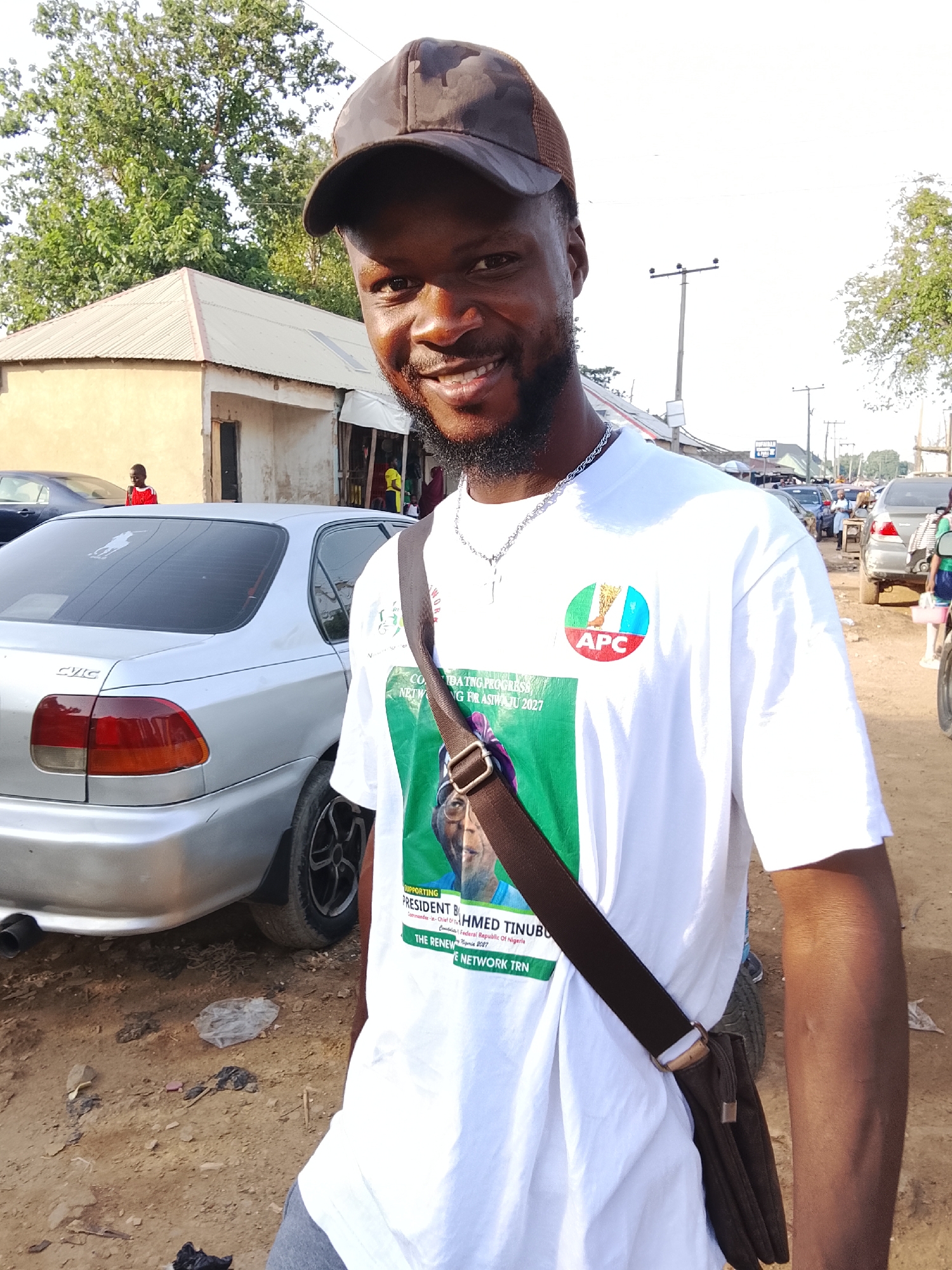 A young man with dark skin, a beard, and a friendly smile poses for a photo outdoors in what appears to be a street in Abuja, Nigeria. He wears a dark camouflage baseball cap, a silver chain necklace, and a white t-shirt, with a brown messenger bag slung across his body. The white t-shirt prominently displays a circular logo with "APC," the acronym for Nigeria's All Progressives Congress political party. Below this, text reads "SUPPORTING PRESIDENT B. AHMED TINUBU," identifying him as the Commander-in-Chief of the Federal Republic of Nigeria. Additional visible text on the shirt includes "NETWORKING FOR ASIWAJU 2027" and "THE RENEW E NETWORK TRN," indicating political advocacy and support for President Bola Ahmed Tinubu.
The background shows a sunlit, clear day. A silver Honda Civic car is parked on the left with "CIVIC" visible on its tail lights and a Polo Ralph Lauren sticker on the rear window. The ground is unpaved dirt with scattered refuse, suggesting a somewhat informal urban or semi-urban setting. In the distance, several low-rise buildings with corrugated metal roofs, utility poles, and power lines are visible, along with indistinct figures of other people and vehicles. The overall scene captures a moment of political expression within a bustling street environment.