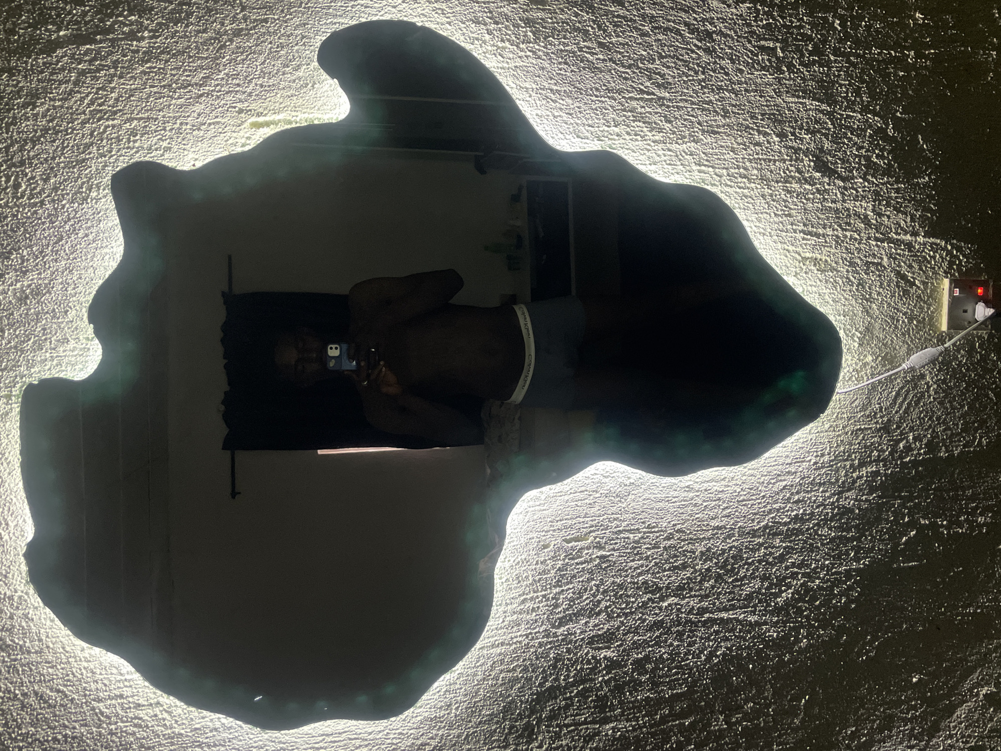 A man is taking a selfie in a mirror shaped like the continent of Africa, which is illuminated with a bright white light outlining its edges. The mirror is mounted on a textured wall, with an electrical outlet and charging cable visible nearby. The background reflected in the mirror shows a glimpse of an interior space. There is no evidence to confirm the location of this image is in Jos, Nigeria.