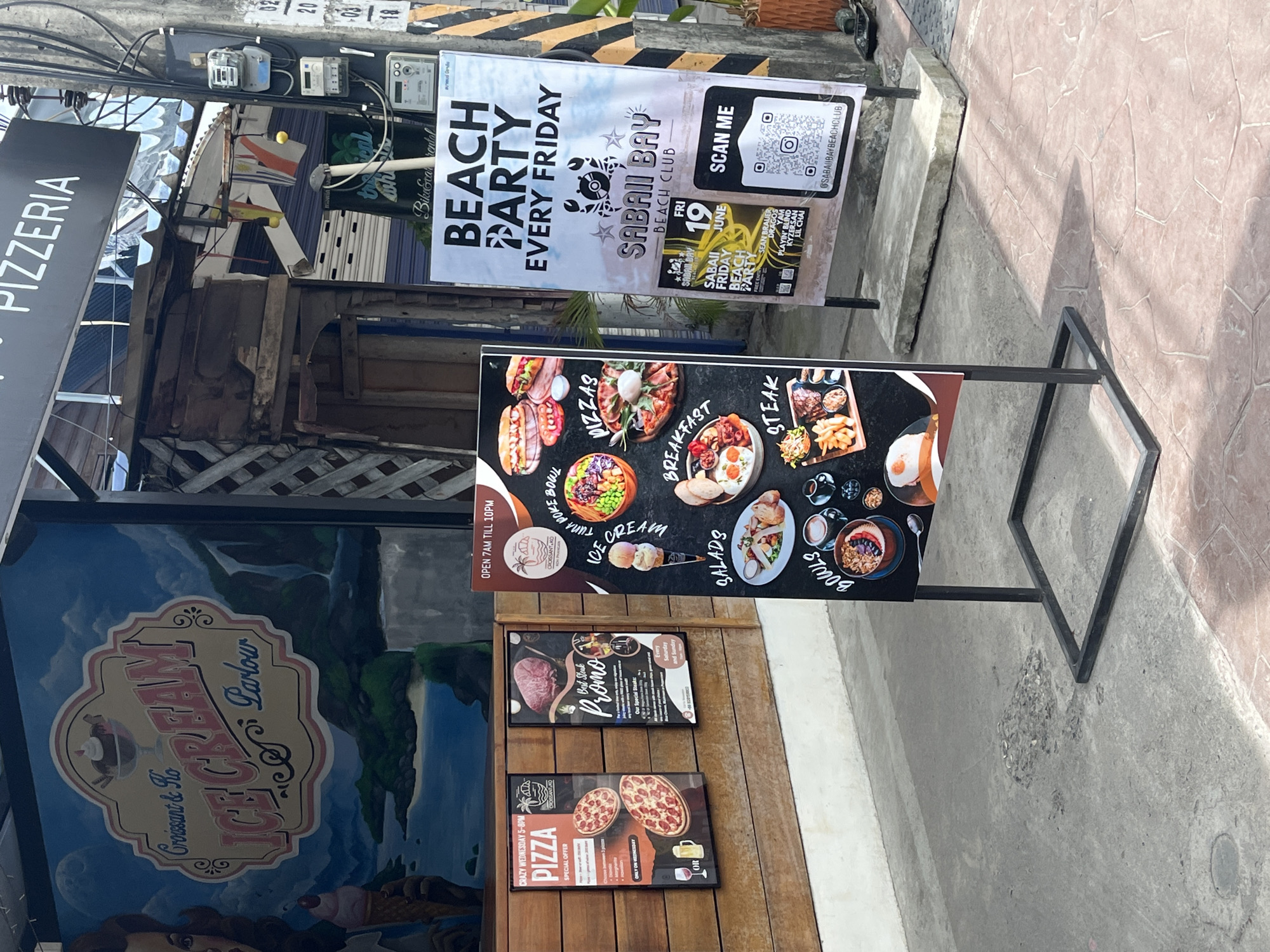 The image shows a street view outside a restaurant in Koh Phangan, Thailand, specifically on the island of Koh Phangan. The building features a mural of an ice cream parlor with the text "Croissant & Co Ice Cream Parlour". To the right of the building, there are several posters. One poster advertises "BEACH PARTY EVERY FRIDAY" at "SABAI BAY BEACH CLUB". Another poster displays food items like "PIZZA" and "Best Steak Promo". A third, more detailed menu poster lists "TUNA POKE BOWL", "ICE CREAM", "PIZZAS", "SALADS", "BREAKFAST", and "STEAK". The ground in front of the establishment is paved with concrete and stone tiles. Electrical meters and wiring are visible on a utility pole. The lighting suggests it is daytime.