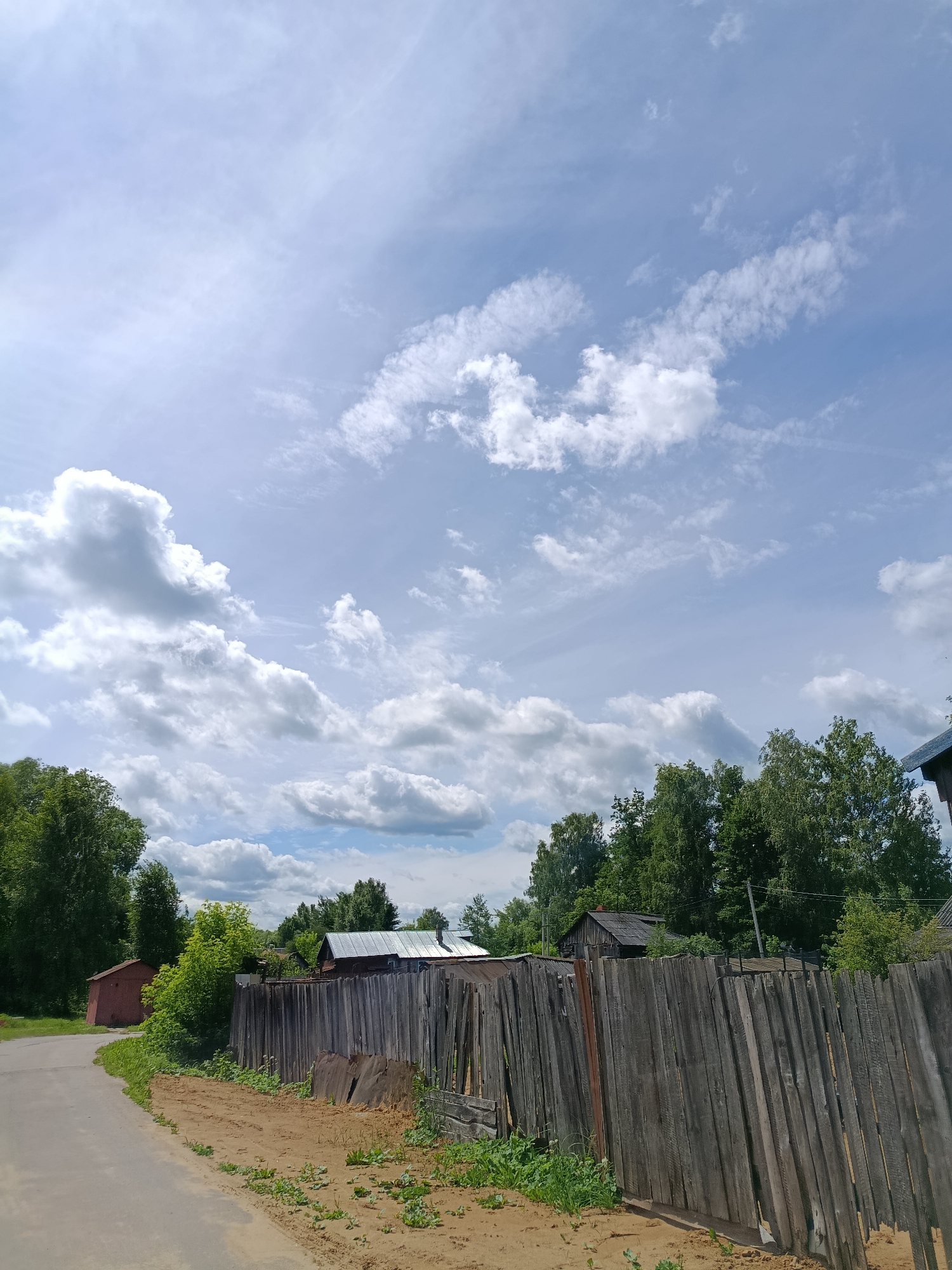 The image depicts a rural scene with a weathered wooden fence in the foreground. Beyond the fence, there are several rustic wooden buildings and dense green trees. A paved road curves to the left, leading towards a small red building. The sky is partly cloudy with cumulus clouds against a pale blue background. The overall impression is of a peaceful, slightly unkempt, rural environment.