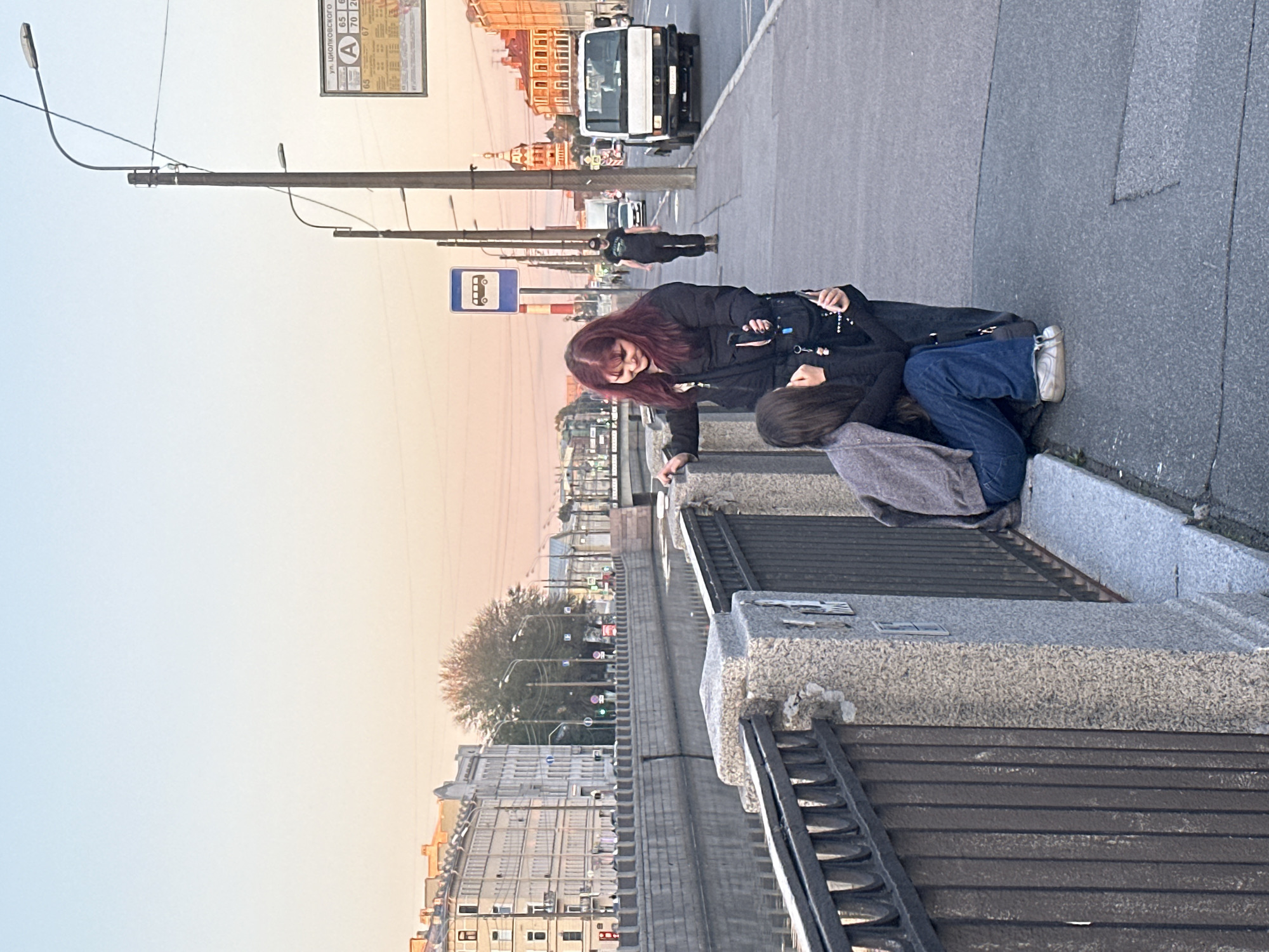 The image depicts a street scene in Saint Petersburg, Russia, likely at dusk, given the soft, pastel hues of the sky. In the foreground, two young individuals are interacting on a sidewalk adjacent to a canal. One person, with long, dark hair, is standing, leaning over a stone embankment with a metal railing. They are wearing dark clothing, including a jacket. The other person, with dark hair and wearing a grey cardigan over dark clothing and blue jeans, is kneeling or sitting by the railing, appearing to be consoled or engaged in an intimate conversation with the standing individual.
The environment suggests an urban setting. Across the canal, a row of multi-story buildings with classical architectural features lines the opposite bank. Trees are visible interspersed among the buildings. On the sidewalk where the individuals are standing, a bus stop sign is present, along with streetlights and other poles. A white truck is parked further down the sidewalk, and other vehicles and a pedestrian are visible in the background, suggesting a moderately busy street. The canal itself is calm, reflecting the muted light. The overall atmosphere is subdued, with the focus on the interaction between the two individuals.