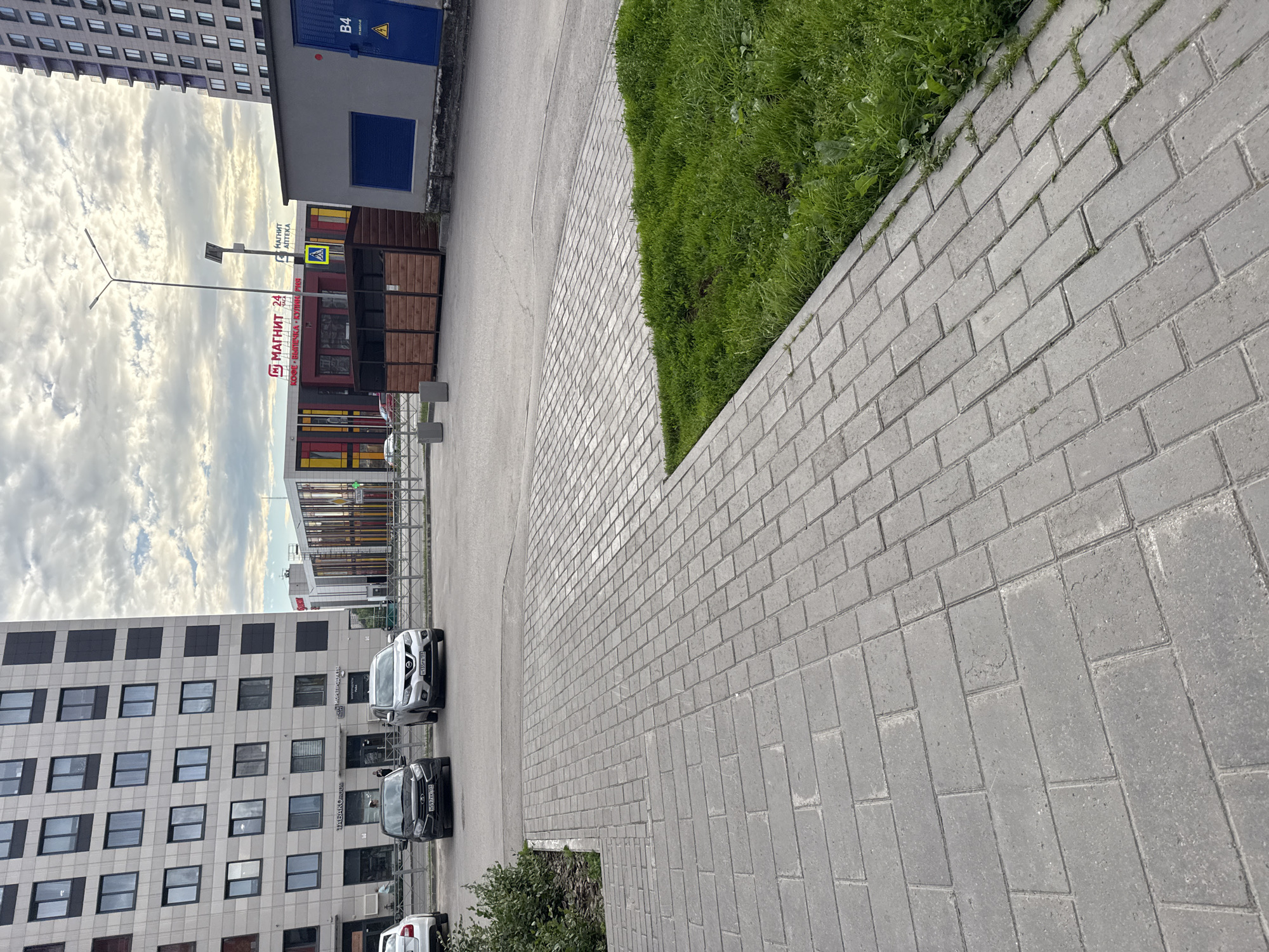 The media file depicts an outdoor scene in Perm, Russia, on a cloudy day. The perspective is from ground level, looking upwards and across a paved area.
In the foreground, a brick-patterned sidewalk made of grey paving stones dominates the right side of the frame. Adjacent to the sidewalk is a strip of vibrant green grass. Further into the scene, a grey asphalt road is visible, bordered by the sidewalk and grass.
On the left side of the image, a modern, multi-story building with a grey facade and numerous dark-framed windows is prominent. The building appears to be an apartment complex or office building. To the right of this building is a smaller, grey structure with a blue door labeled "B4". Next to this structure is a commercial kiosk with wooden paneling and bright signage. Visible text on the kiosk includes "МАГНИТ 24" (Magnit 24), "КОФЕ • ВЫПЕЧКА • КОЗИНАКИ" (Coffee • Pastries • Kozinaki), and "МАГНИТ АПТЕКА" (Magnit Pharmacy). A pedestrian crossing sign with a yellow diamond and a black silhouette of a person is also visible near the kiosk.
Several cars are parked on the asphalt road. A silver sedan is parked closest to the buildings, followed by a dark-colored sedan behind it, and partially visible, a white car at the very end.
The sky is filled with grey, textured clouds, suggesting overcast weather. The overall impression is that of an urban or suburban environment, likely a residential or commercial area, during the daytime. There are no people visible in the media file.