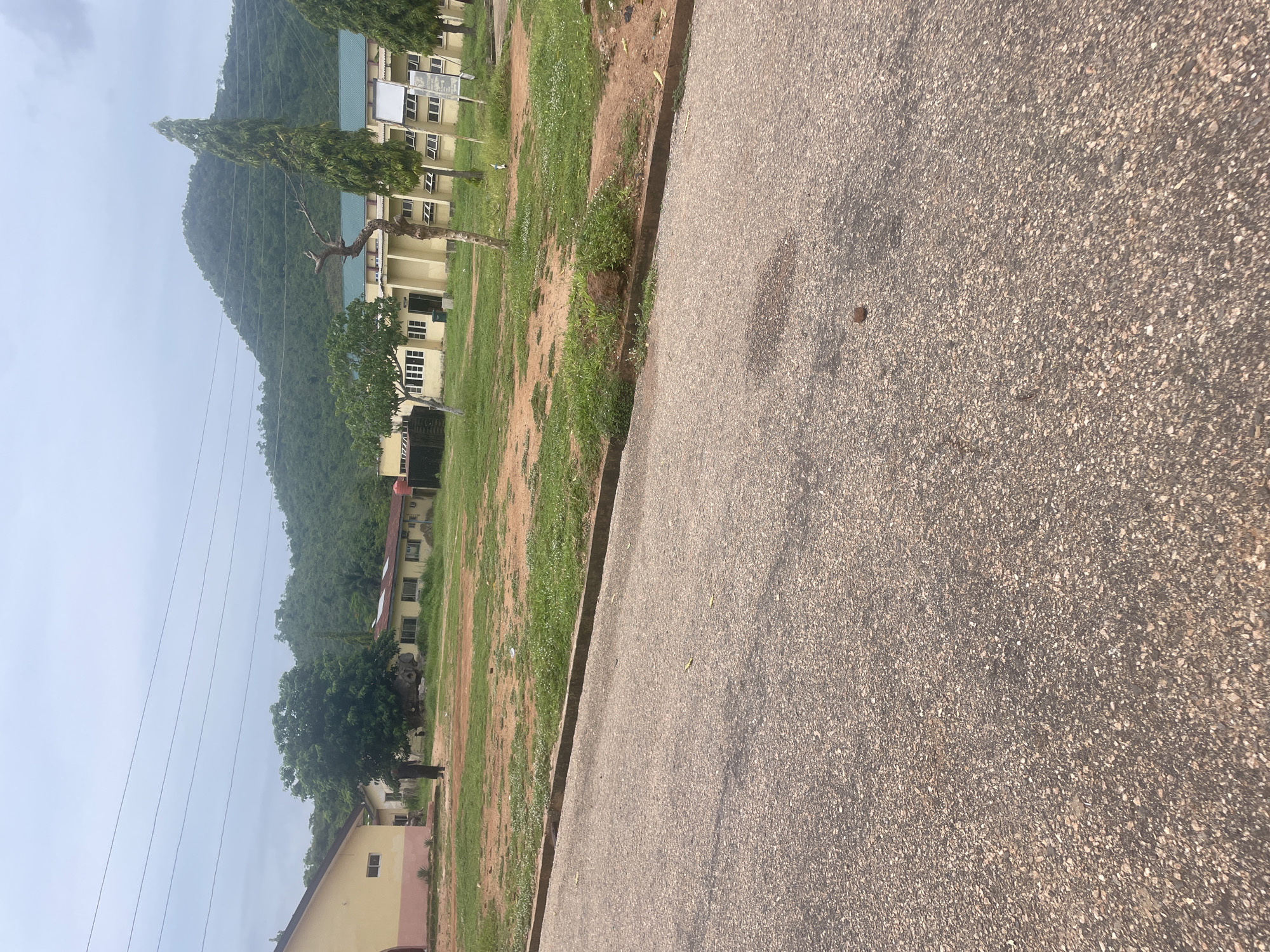 The image shows a view of a street in Lokoja, Nigeria, with a building complex in the background. The foreground is dominated by an asphalt road, with a curb and a patch of dry grass next to it. In the middle ground, there is a large open area with sparse green grass and patches of reddish-brown earth. A few trees are scattered across this area, and a person is standing near one of them.
The background features a cluster of light yellow buildings with green roofs, likely part of an institutional complex. The architecture suggests a functional, perhaps educational or administrative, setting. Behind the buildings, a lush, green, tree-covered hill rises steeply, characteristic of the topography of Lokoja, which is situated at the confluence of the Niger and Benue rivers. The sky is overcast, indicating a cloudy day. The overall scene depicts an outdoor environment in a Nigerian city, with a mix of natural landscape and man-made structures.