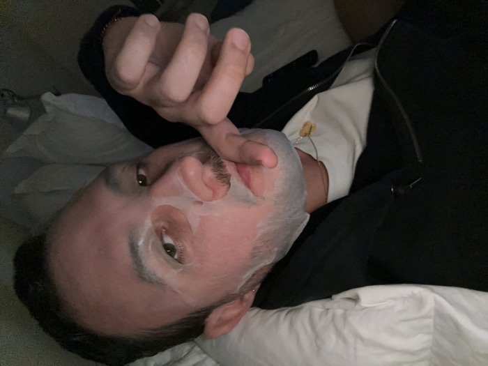 An adult male is depicted lying in an indoor setting. He has short dark hair, brown eyes, and a mustache. A translucent white sheet-style face mask covers his cheeks, nose, and forehead, with cutouts allowing his eyes and mouth to be visible. His right hand, partially visible, is raised with fingers touching the mask near his mouth and chin, suggesting an action of application or adjustment. He is wearing a white t-shirt underneath an unzipped black jacket or hoodie. A thin chain necklace with a small, light-colored, possibly amber-like pendant rests on the white t-shirt. The individual is resting on white bedding, likely a pillow and duvet. The surrounding environment is dark and lacks distinct features, rendering a precise location within the city of London, United Kingdom, or any other specific geographical area, indeterminable from the visual evidence.