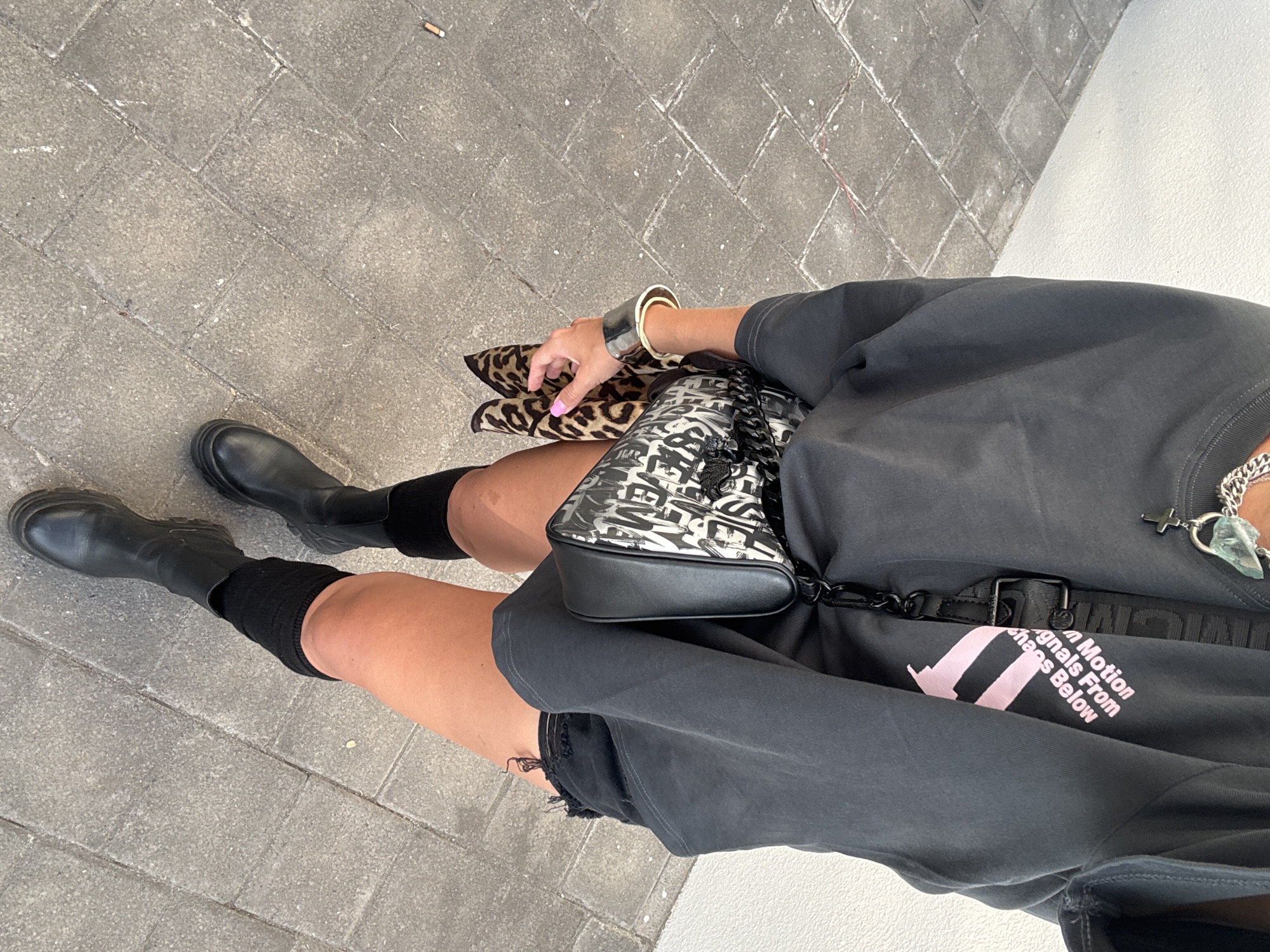 A full-body shot from a slightly high angle shows a person from the chest down standing on a paved walkway. The person is wearing black ripped shorts, a dark gray sweatshirt, black knee-high socks, and black chunky boots. A black purse with a white graphic print is slung across their body, and a leopard-print scarf is tucked into the side of the purse. A thick metal bangle is worn on the left wrist. The background is a white wall and a gray paved surface, suggesting an outdoor urban setting. The lighting is bright and appears to be daylight.