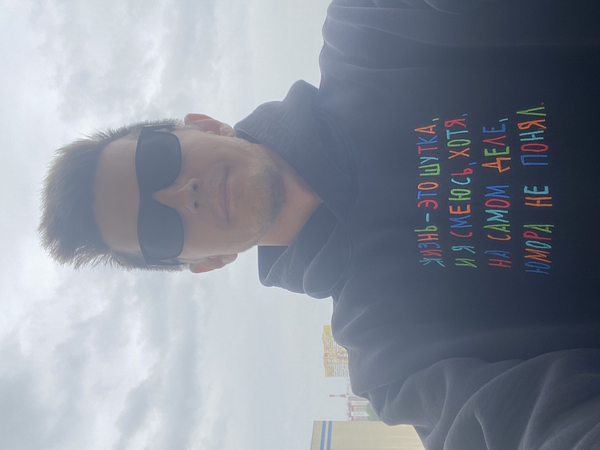 This is a selfie of a person wearing sunglasses and a dark hoodie. The hoodie has colorful text embroidered on it, which appears to be in Russian. The background shows a cloudy sky and a cityscape, with buildings visible. The image may have been taken in Rozhdestveno, Russia, based on the provided prompt. The person is looking directly at the camera, and their face is partially obscured by sunglasses. The focus is on the person and the text on their hoodie.