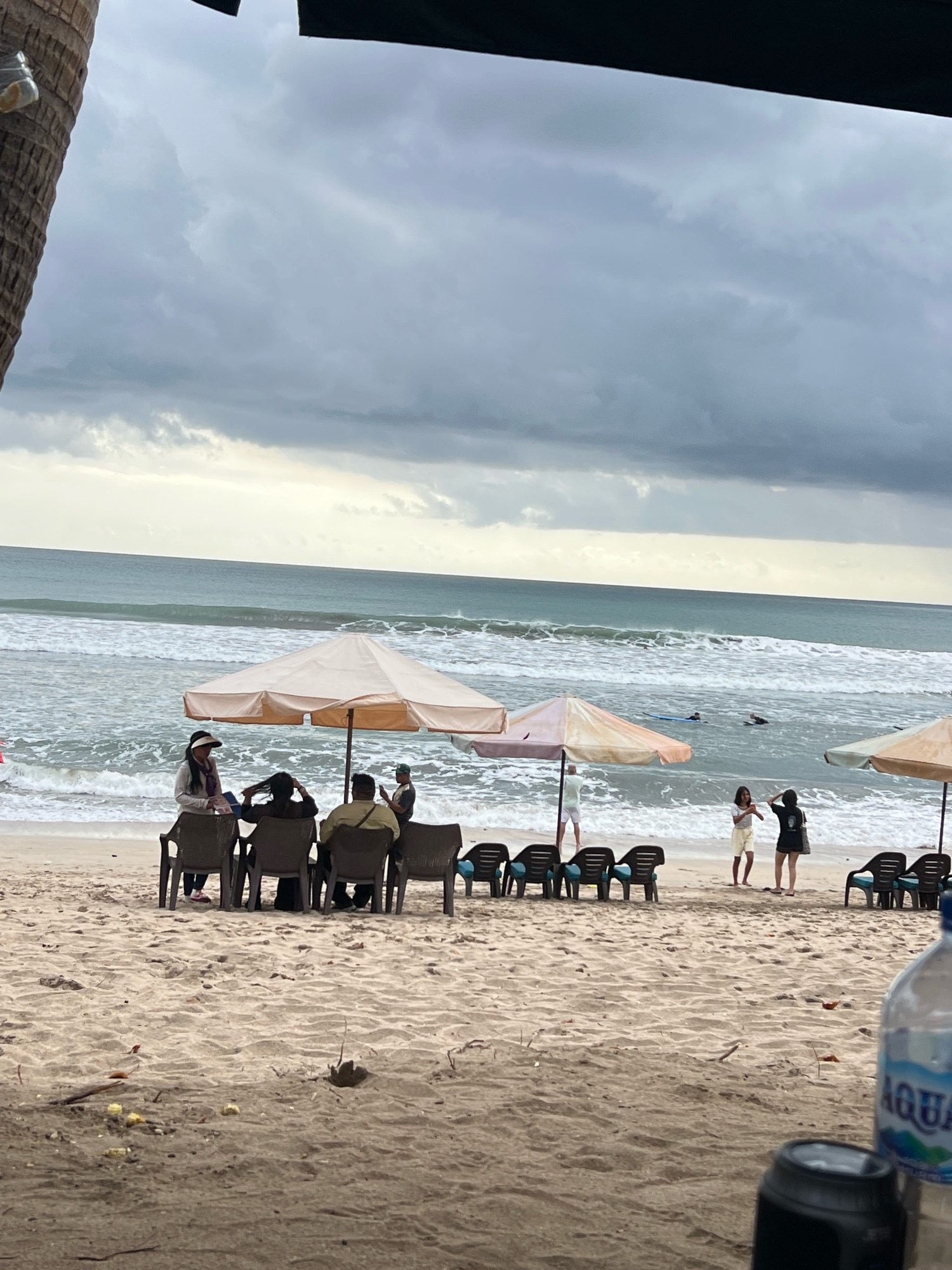 A cloudy day at Kuta Beach, Indonesia. Several people are relaxing on the sand under umbrellas, while others are in the ocean, some with surfboards. In the foreground, a bottle of Aqua water and a camera lens are visible, suggesting the viewpoint is from a table. The waves are moderate, and the sky is overcast with dark clouds.
