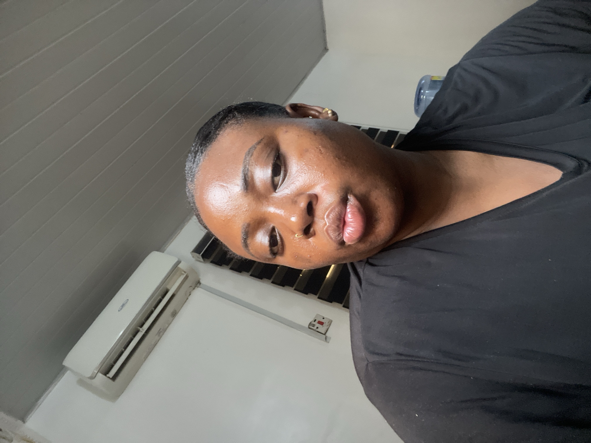 A medium-close shot shows a Black woman with smooth, dark skin, wearing a black top. She has dark hair pulled back, a nose ring, and earrings. The background includes a white wall, an air conditioning unit, and a water dispenser with a large blue bottle. The location appears to be indoors. The city is not directly discernible from the image, so it is impossible to confirm the location as Abuja, Nigeria from visual information alone.
