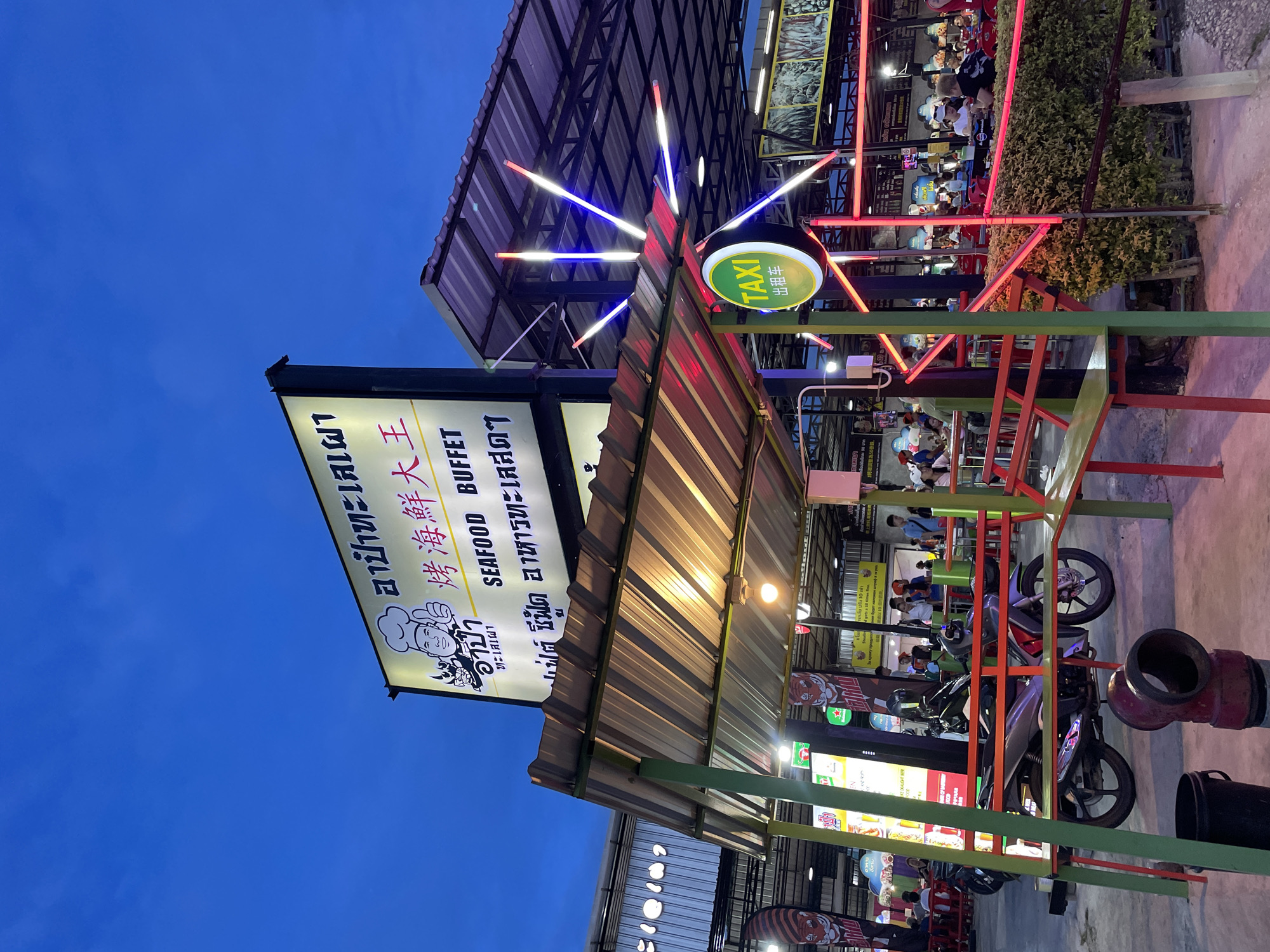 The image depicts an outdoor dining area at night in Pattaya City, Thailand. A prominent illuminated sign with Thai and Chinese characters and an image of a chef is visible, advertising "SEAFOOD BUFFET" and "烤海鮮大王" (King of Grilled Seafood). Red, blue, and white neon lights form a starburst pattern above a green and yellow "TAXI" sign. The seating area features tables and chairs, some occupied by patrons, and is illuminated by overhead lights and the colorful neon accents. A motorcycle is parked in the foreground, partially covered by a structure with a corrugated metal roof supported by green posts. The atmosphere suggests a lively, casual dining establishment.