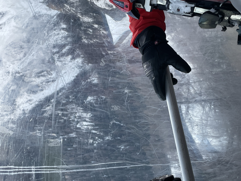 The image presents a close-up, first-person perspective from within what appears to be a ski lift cabin, looking out through a heavily scratched, transparent window. In the foreground, a person's left hand, covered by a black winter glove with a red cuff, is seen gripping a white or light grey ski pole. The brand name "REUSCH" is visible on the glove. Above the hand, part of a ski binding, colored red and metallic, is visible, suggesting the presence of a ski.
The background, viewed through the scuffed window, reveals a snowy, forested mountain landscape. Snow-covered evergreen trees are discernible, indicating winter conditions. The numerous scratches on the transparent surface create a textured, somewhat distorted view of the outside world. The lighting suggests it is daytime. The scene depicts an individual engaged in or traversing a ski area, consistent with a location like Courmayeur, Italy. The activity taking place is travel on a ski lift during a snowy winter day.