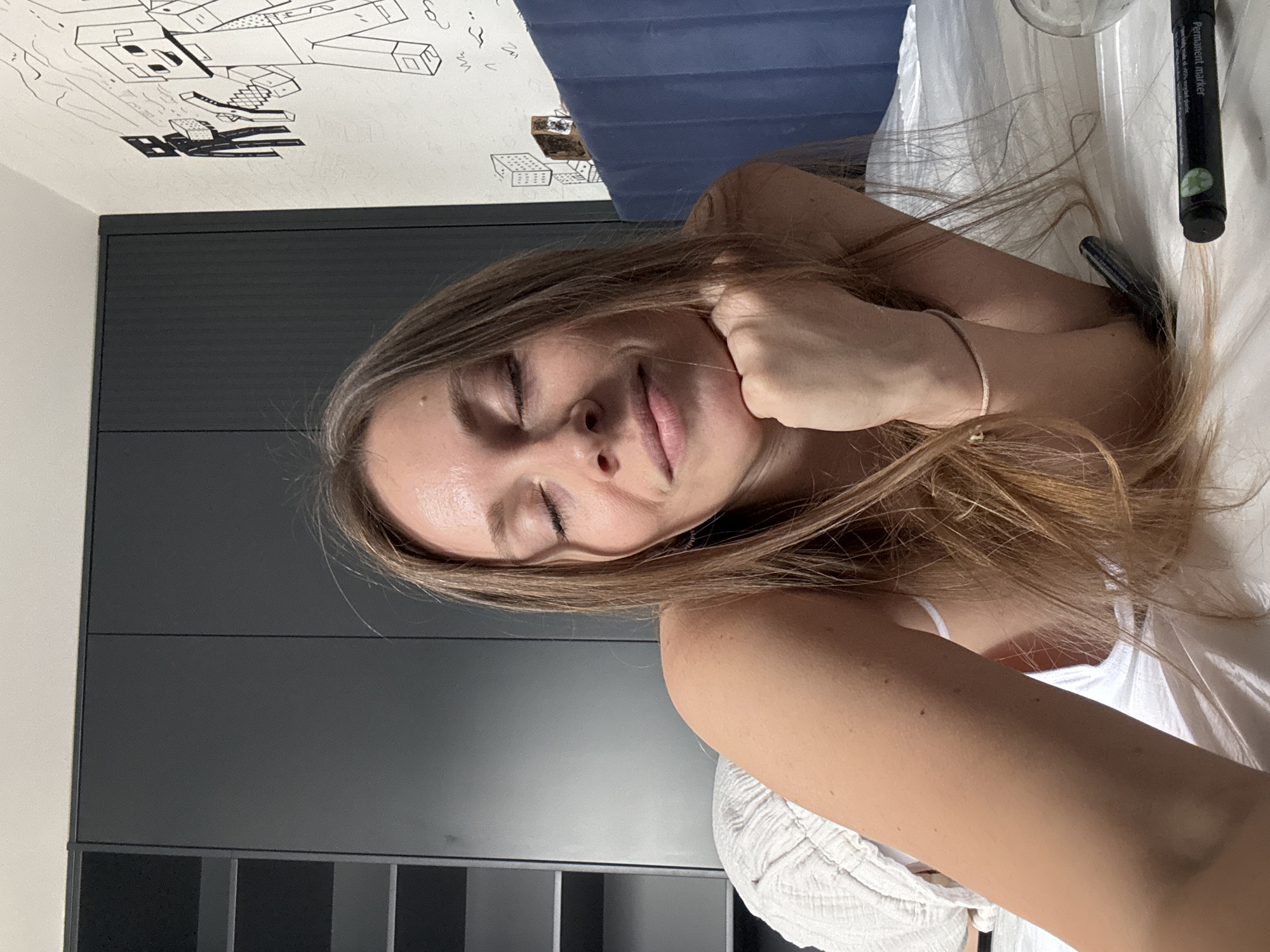 A young adult female lies supine with her eyes closed and hand supporting her head. She is wearing a white top and a thin bracelet. Next to her head is a black permanent marker. The backdrop reveals a grey paneled wall and a ceiling with rudimentary Minecraft-inspired drawings. The location might be a residential room in Budva, Montenegro.