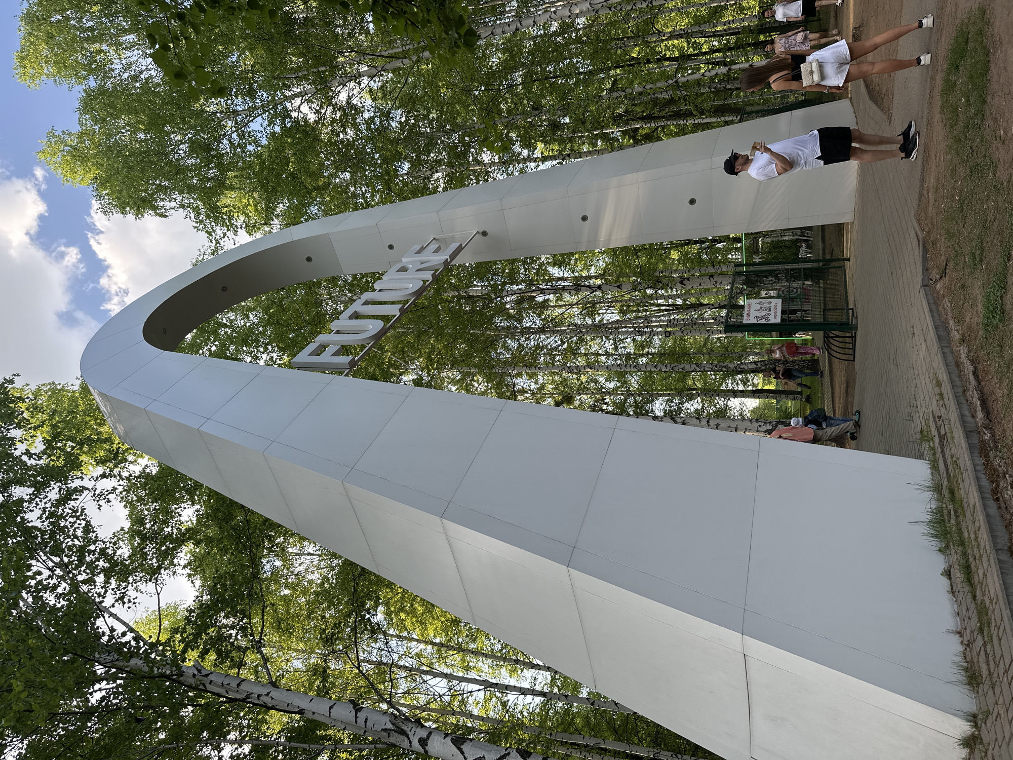 A white arch-shaped structure with the word "IZHEVSK" spans an entryway to a park area in Izhevsk, Russia. The arch is made of a smooth, white material and is segmented into panels. Trees with green leaves are visible in the background, some with white bark.
Several people are present in the scene. One person, wearing a white shirt, black shorts, and a baseball cap, stands under the archway, appearing to be talking on the phone or looking at their phone. Two other people, dressed in summer clothing, are seen in the background, possibly walking or standing. There are other individuals present on the other side of the arch, sitting on or near benches and resting.
The ground is covered in dirt and grass with a paved walkway that leads under the archway. A metal gate and sign can be seen under the arch, indicating the entrance to the park. The sky is partly cloudy.