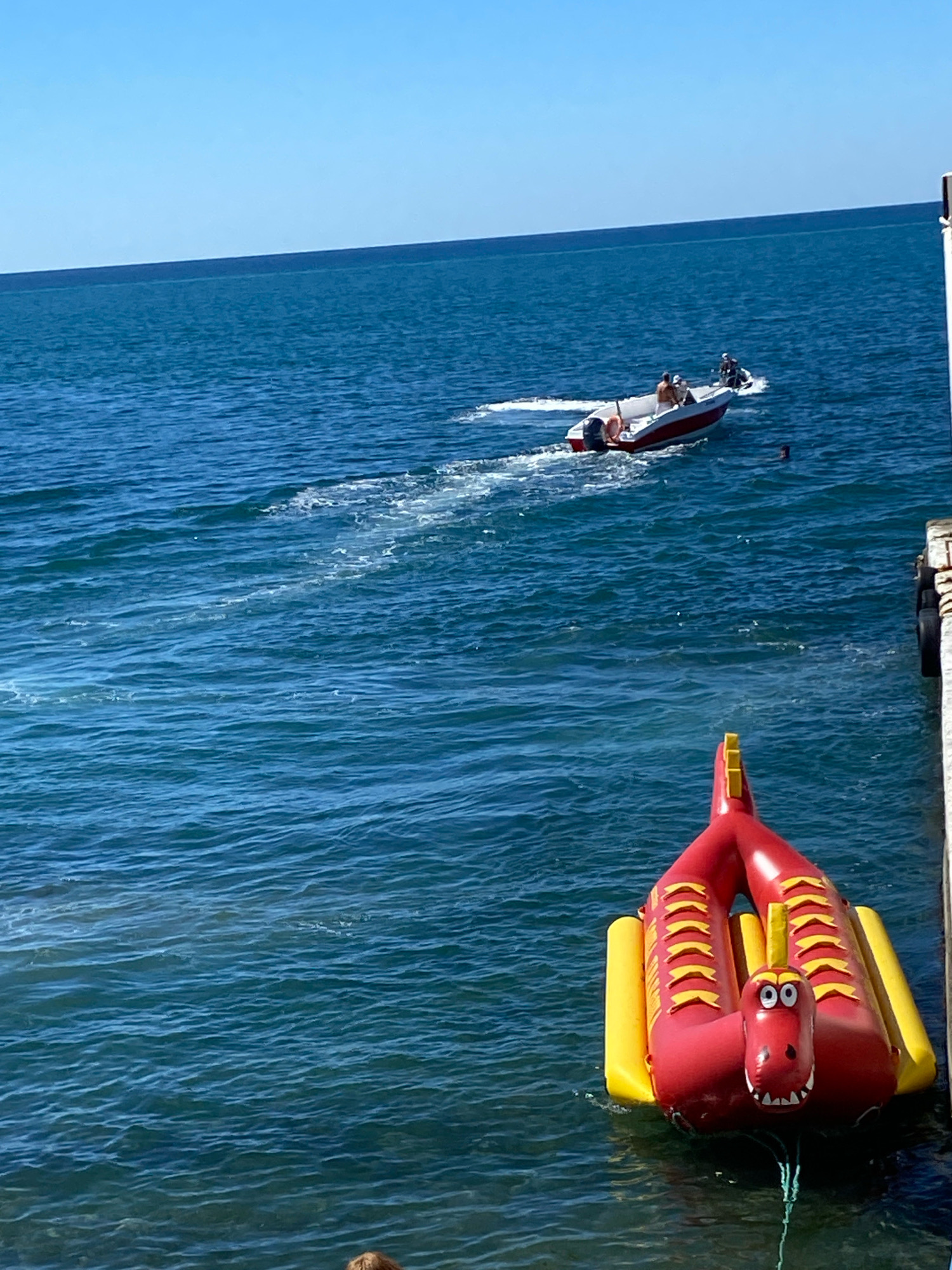 The image depicts a clear blue sky over a calm, dark blue sea, likely the Black Sea off the coast of Sochi, Russia. In the foreground, a red and yellow inflatable dragon-shaped toy floats on the water, tethered by a rope. To the right, a portion of a pier or dock with white vertical supports and black fenders is visible. In the mid-ground, a small white and red motorboat is cutting through the water, leaving a white wake. Several people are visible in and around the boat, with two appearing to be passengers and one possibly operating a jet ski behind the boat. Another individual is swimming in the sea between the inflatable toy and the motorboat. The overall scene suggests a recreational day at the seaside.