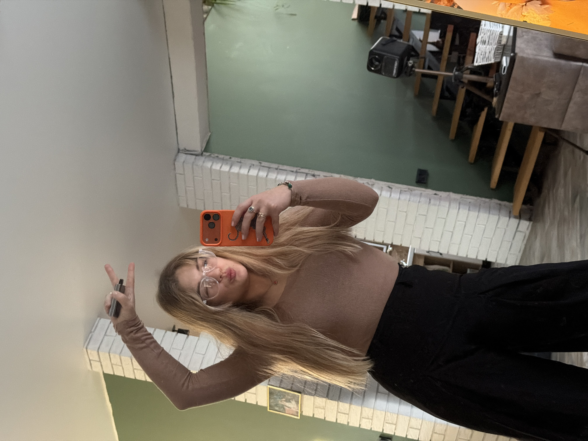 A young woman with long, straight blonde hair and clear-rimmed glasses takes a mirror selfie in an indoor setting. She wears a light brown long-sleeved top and black pants. With her right hand, she holds an orange smartphone featuring a triple-camera setup and a distinct squiggly line pattern on its case. Her left hand forms a peace sign, holding a silver or metallic vaping device between her index and middle fingers. She has multiple rings on her fingers and a bracelet on her left wrist, and her expression is a slight pout or "duck face," conveying a playful mood.
The background, visible in the mirror's reflection, reveals a domestic interior. A prominent feature is a white brick structure, possibly a fireplace or decorative wall, against which a pale green wall is visible. Above the brickwork, a small framed picture hangs. Further into the room, furniture is reflected, including a table with wooden legs and what appears to be a dark-toned sofa or chair. A black GoPro camera, mounted on a stand or tripod, is also visible. The lighting appears soft and ambient, suggesting daytime indoors.