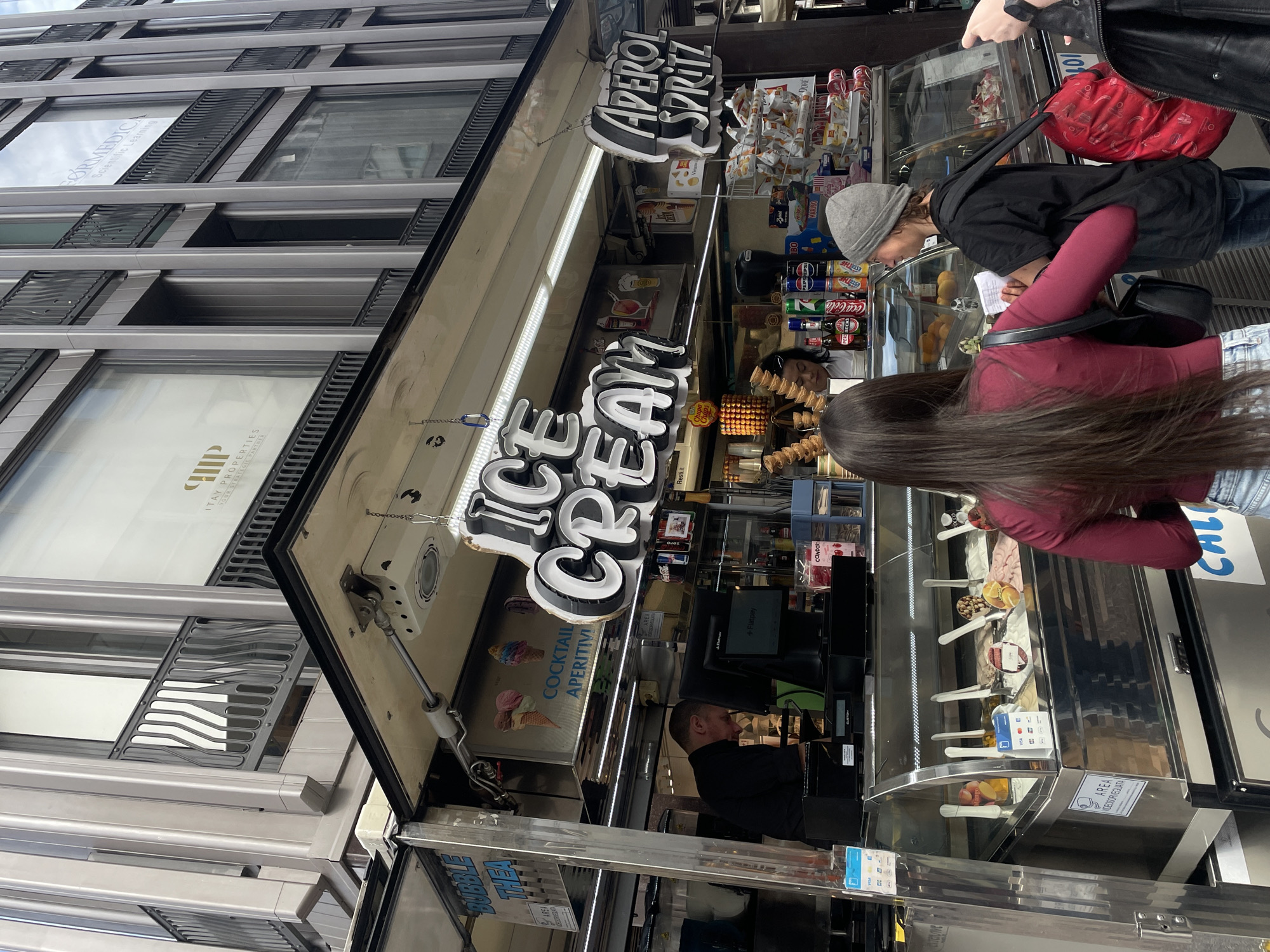 Here is a detailed description of the image:
**Scene:** The image depicts an outdoor ice cream and aperitif stand in what appears to be a city center. Customers are browsing the offerings at the stand.
**Location:** The location can be reasonably inferred to be in Milan, Italy due to the Italian wording of "COCKTAIL APERITIVI", "APEROL SPRITZ" and some signs in Italian "AREA VIDEOSORVEGLATA" and "ITAY PROPERTIES". Also there are some other Italian brands such as "Resti.it" visible on the scene.
**Subjects:**
* Customers: There are at least two customers visible, a person with long brown hair wearing a red shirt, and another person with a grey beanie wearing a black jacket. They are looking at the items in the display case of the ice cream stand.
* Vendor: A man is standing inside the ice cream stand, presumably the vendor.
**Objects:**
* Ice Cream Stand: The stand has a sign that says "ICE CREAM" in large, illuminated letters. Above it, another sign reads "APEROL SPRITZ" and the stand also features a smaller sign for "COCKTAIL APERITIVI". There is a display case with various ice cream flavors and a selection of drinks and snacks behind the counter. Also, in the side of the stand there is another sign of "BUBBLE THEA".
* Building: The stand is located in front of a modern building with horizontal window designs. There is a sign visible that says "FØRMEDICA" on the building's windows.
* Drinks and Snacks: There are drinks like Coca-Cola, Pepsi, ESTATHÉ, some snack brands like Chupa Chups, Haribo and bags of snacks behind the counter.
**Actions/Interactions:**
* Browsing: Customers are actively looking at the ice cream and aperitif offerings, possibly deciding on a purchase.
* Serving: The vendor is likely preparing to serve or is in the process of serving customers.
**Environment:**
* The scene is outdoors, likely on a busy street.
* The presence of the building suggests an urban environment.
**Other details:**
* Other signs: The presence of a small sign "Flatpay" and another sign stating "AREA VIDEOSORVEGLATA" that implies video surveillance.