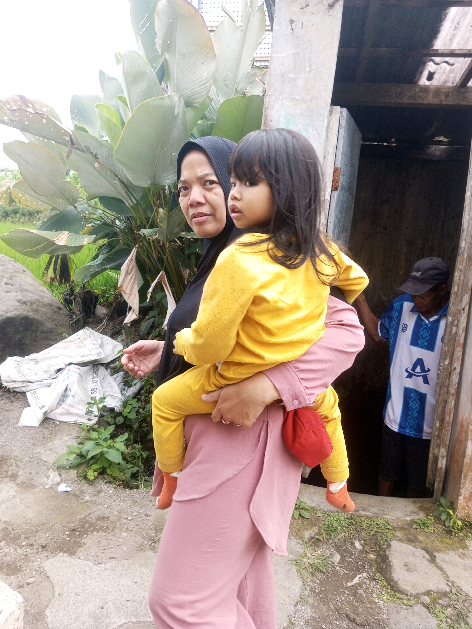 A medium shot shows a woman wearing a black hijab and a pink abaya carrying a young child. The child, dressed in a yellow top and pink pants with orange socks, is positioned on the woman's left hip, facing right. Her right arm is around the child's torso. The woman's expression is directed towards the viewer. In the background, a man wearing a blue and white striped shirt and a black cap is partially visible standing in a doorway. The environment appears to be outdoors with large green leaves and some sacks on the ground. The scene is set near a dilapidated structure with a wooden door. The overall impression is of a casual, everyday moment in what appears to be a rural or semi-rural setting in Krasak, Indonesia.