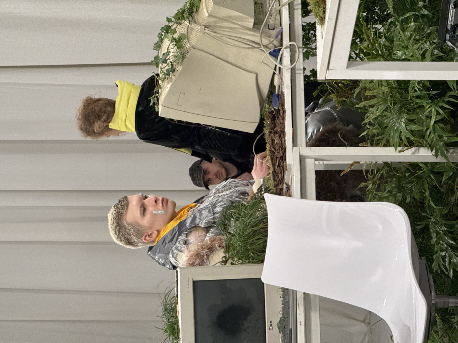 This image depicts an indoor scene, likely an exhibition or curated event, taking place in Milan, Italy. Three individuals are visible amidst a setting characterized by a blend of obsolete technology and overgrown plant life.
In the left foreground, a young male with short, bleached blonde hair and a metallic silver patch on his right cheekbone wears a yellow high-neck garment beneath a textured, multi-toned jacket, possibly featuring faux fur or distressed fabric and a subtle camouflage pattern. He gazes intently towards the right, out of frame.
Behind him and to the center, a male wearing a dark baseball cap and stubble looks directly at the camera with a neutral expression, partially obscured by the first individual and desk elements.
To the right, a third person, seen from behind, has curly reddish-brown hair and wears a dark jacket with a prominent, bright yellow, puffed collar or scarf. This individual is also oriented towards the right, out of frame.
The scene is furnished with white-framed desks. On the left desk, an old, beige cathode ray tube (CRT) monitor labeled "Low Radiation" is visible, partially integrated with small green plants. The right desk features another beige CRT monitor, tilted and heavily adorned with green ivy vines and loose electronic wires, including a blue VGA cable. The desk surfaces are covered with dark brown mulch or wood chips and interspersed with various green plants and grasses. The area beneath the desks is also filled with dense foliage, primarily ferns. An unoccupied white, modern, ergonomically-shaped swivel chair is positioned in the left foreground. The background consists of light-colored, plain walls or drapes.