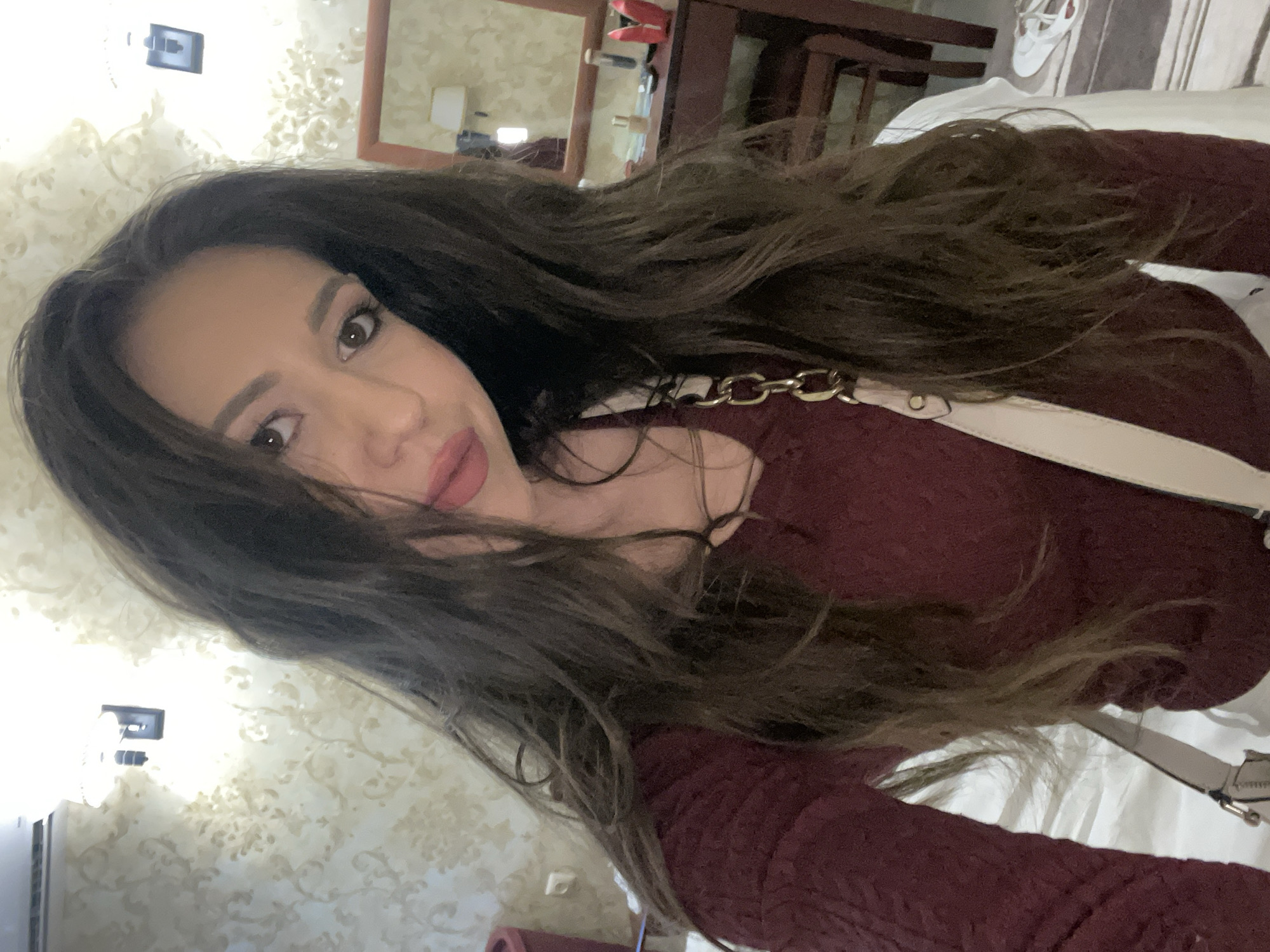 A woman with long, dark hair, wearing a maroon sweater and a light-colored purse with a gold chain strap, is the primary subject of this indoor photograph. She is facing the camera with a slight smile. The background includes a patterned wall and furniture, suggesting a hotel or home setting. The location might be Starozagorski bani, Bulgaria.