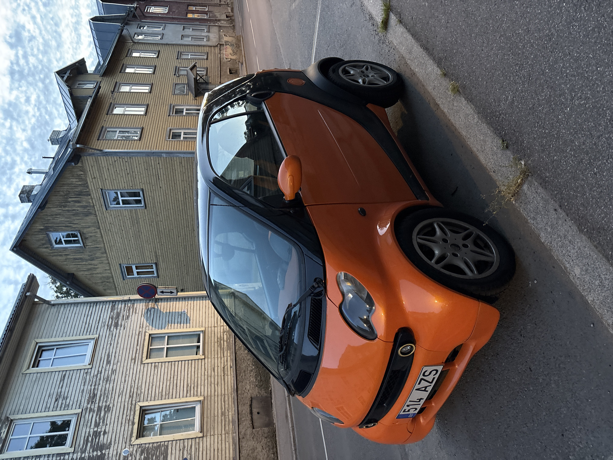 The image shows a small orange and black Smart car parked on the side of a street in Tallinn, Estonia. The car, with license plate "514 AZS", is positioned at an angle to the viewer, with its front-left quarter most prominent. The street appears to be paved with asphalt, and a concrete curb with some sparse vegetation is visible along the right edge of the image.
In the background, a row of old wooden buildings lines the street. The building immediately behind the car is painted a light yellow and has a gabled roof. To its left, another building is visible, painted a weathered greyish-beige, also with wooden siding and a gabled roof. Further down the street, more buildings with similar architectural styles can be seen, some appearing to be multi-story. A traffic sign with a red circle and a blue horizontal bar, indicating no parking or stopping, is visible on a pole between the two closest buildings. The sky above is partly cloudy with a mix of blue sky and white clouds. The lighting suggests it is either early morning or late afternoon, with the sun casting soft shadows. No people are visible in the image.