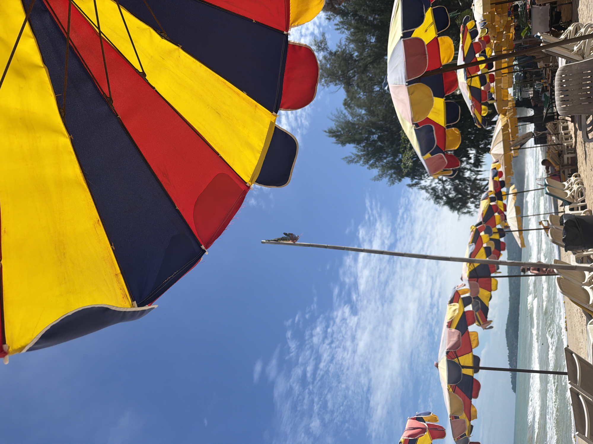 The image depicts a beach scene in Choeng Thale, Thailand, on a sunny day with a clear blue sky and scattered clouds. Several beach umbrellas with red, yellow, and blue panels are visible, with one large umbrella dominating the foreground. The umbrellas are set up on a sandy beach, with rows of empty lounge chairs in the middle ground. The ocean is in the background, with gentle waves rolling onto the shore. In the distance, a tree-covered hill or island can be seen. A tall, thin flagpole with a small flag is also present. A few people are visible, some sitting on lounge chairs and one person standing near the water. The overall atmosphere is one of a relaxed beach day.