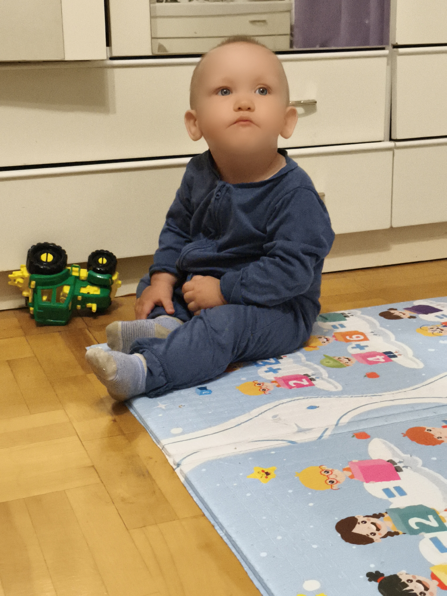 A light-skinned baby with blue eyes and light brown hair is sitting on a light blue playmat on a wooden floor. The baby is wearing a blue zippered jumpsuit and grey socks. The baby is looking up and to the right. A green and yellow toy truck with black wheels is on the floor to the left of the baby. Behind the baby is a white cabinet with drawers and a purple curtain. The scene is indoors.