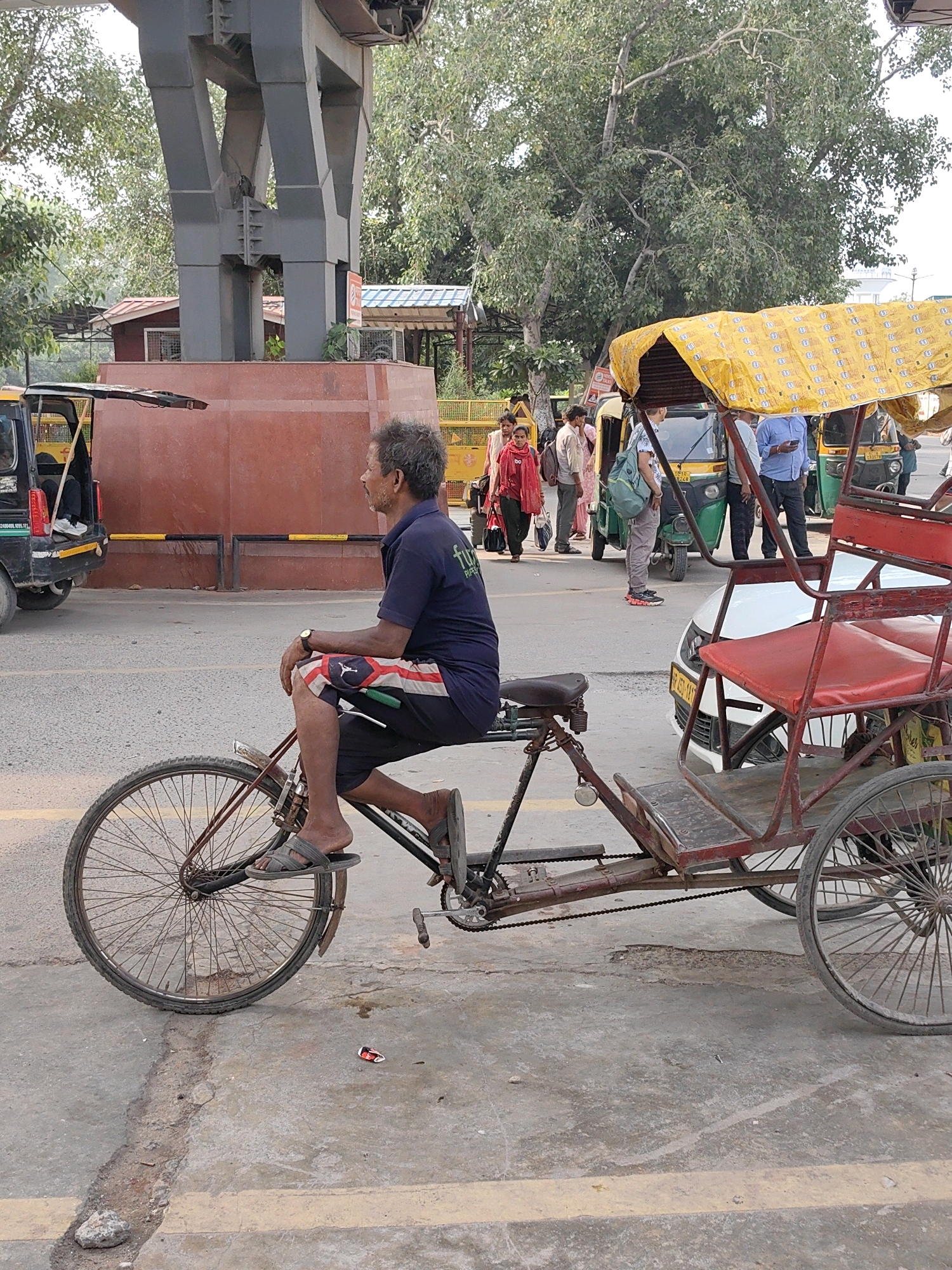 The image captures a scene in Delhi, India, likely near a transportation hub or busy street, as evidenced by the presence of auto-rickshaws and a cycle rickshaw. In the foreground, a man with grey, unkempt hair is seated on a cycle rickshaw, facing right. He is wearing a dark blue t-shirt with a logo and black shorts with red and white accents. His right foot rests on the pedal, and his left foot is on the footrest. He appears to be waiting or resting, with his left hand resting on his knee.
The cycle rickshaw itself is a prominent subject, featuring a red metal frame and a wooden passenger seat. A yellow canopy, patterned with what appear to be small circular logos, is attached to the back, providing shade. The rickshaw has two large spoked wheels at the rear and one at the front.
In the background, several auto-rickshaws are visible, suggesting a public transport area. One auto-rickshaw, partially obscured, is black and has its rear door open. Other auto-rickshaws, typically found in India, are green and yellow. A white car is parked behind the cycle rickshaw.
A group of people are standing and walking near the auto-rickshaws, indicating a bustling environment. One woman in the background is wearing a red garment. The backdrop includes trees with lush green foliage and a large concrete structure, possibly part of an overpass or a building. The ground is paved with asphalt and concrete, with some visible cracks and yellow painted lines. The overall atmosphere suggests a typical day in a busy Indian city, with a focus on local transportation and street life.