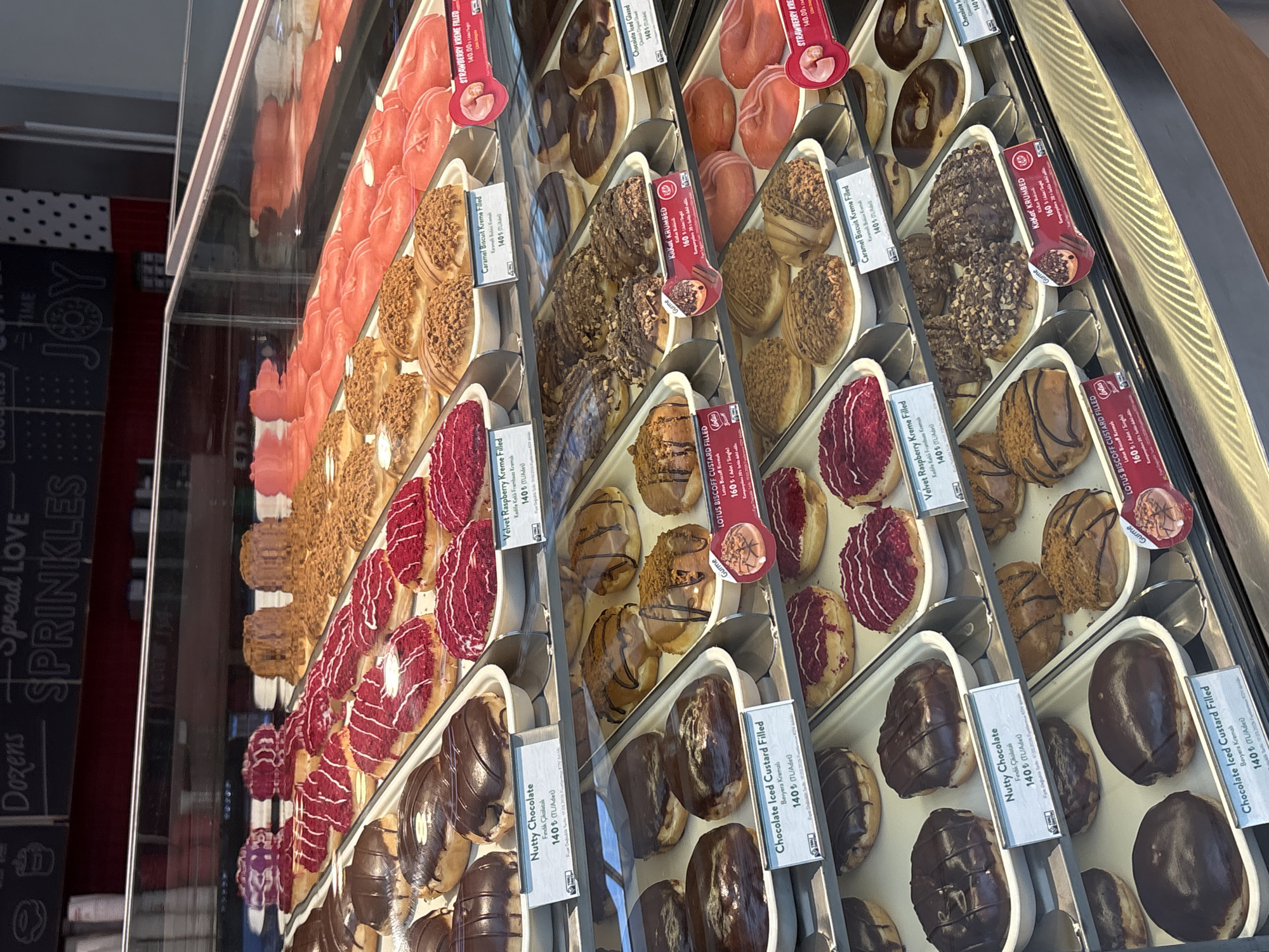 This image captures a multi-tiered glass display case filled with a diverse assortment of donuts, likely within a Krispy Kreme or similar donut shop located in Şişli, Turkey.
The display features multiple rows and columns of individually presented donuts. The top shelves showcase an array of red-glazed and white-drizzled donuts, alongside rows of light brown, possibly caramel or biscuit-topped varieties, and a prominent section of bright pink or red strawberry-flavored donuts. Visible labels indicate varieties such as "Strawberry Kreme Filled" priced at 140.00 TL and "Velvet Raspberry Kreme Filled" also at 140 TL.
The middle shelves exhibit various chocolate-glazed donuts, some plain dark chocolate, others with nut toppings. Specific labels identify "Nutty Chocolate" and "Chocolate Iced Custard Filled" donuts, each priced at 140 TL. Another section displays donuts with a brown, crumbly topping, including "Caramel Biscuit Kreme Filled" (140 TL) and "KitKat Krumbed" (160 TL), featuring a small graphic of a KitKat donut. A "Lotus Biscoff Custard Filled" donut, priced at 160 TL, is also visible.
The bottom shelves continue the theme with more dark chocolate-glazed and custard-filled options, alongside raspberry-drizzled and crumb-topped donuts. The donuts are arranged on white paper trays or liners, and the display case is well-lit, highlighting the colors and textures of the pastries.
In the background, a dark, chalkboard-style wall is visible with white text that reads "Dozens," "Spread Love," "Sprinkles," "Coffee," and "Joy," suggesting a positive and inviting atmosphere common to cafes or bakeries. No individuals are visible in the scene, focusing entirely on the donut selection and display. The currency indicated on the price tags (TL - Turkish Lira) confirms the location within Turkey, specifically Şişli as requested.