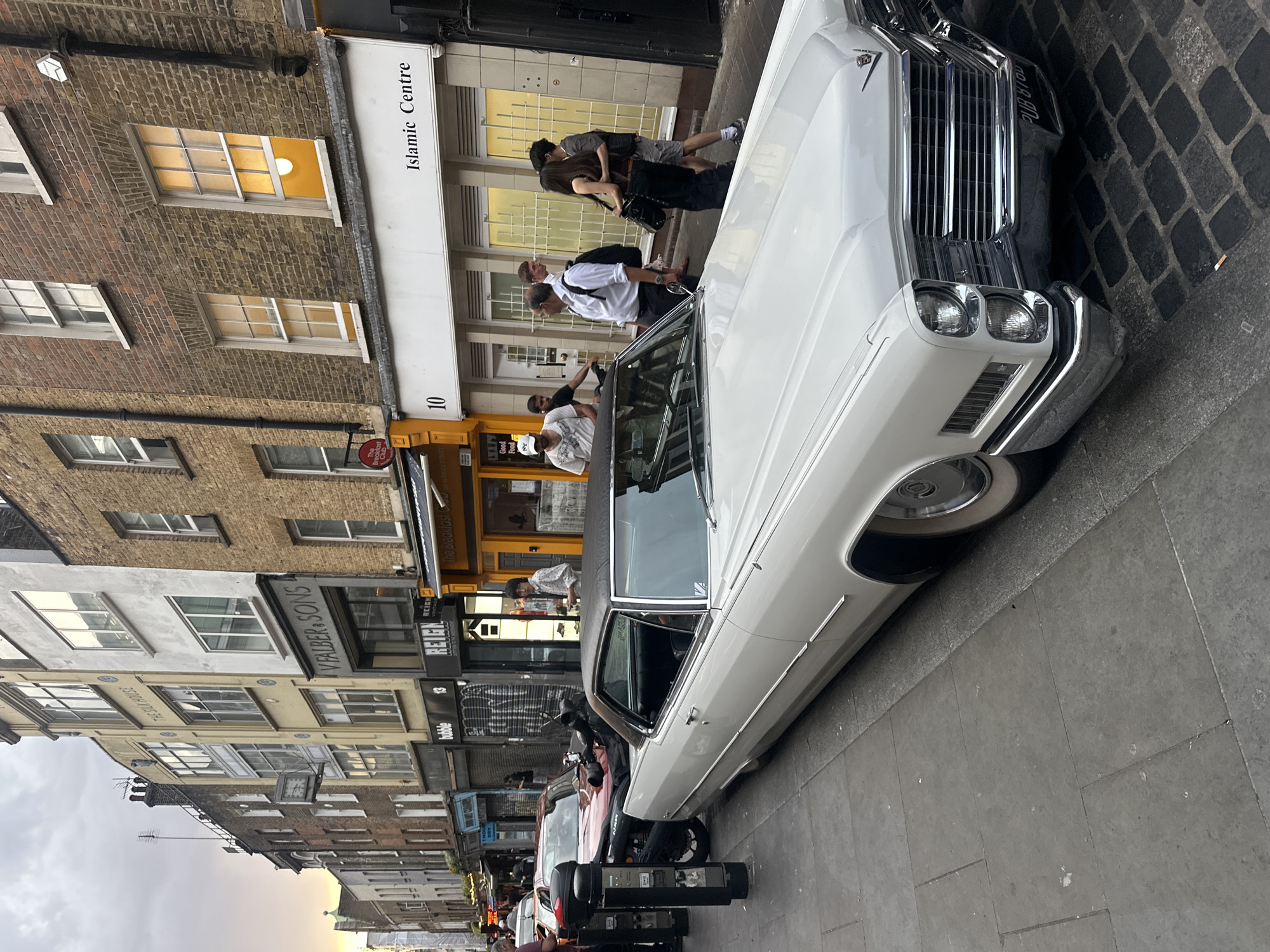 This image depicts a street scene in London, United Kingdom, featuring a vintage white Cadillac parked on the side of the road. Several people are visible, walking and interacting near storefronts. The street includes establishments such as "The Breakfast Club" and an "Islamic Centre," among other shops with signs like "VFALBER & SONS" and "bubble". The architecture includes brick buildings and stone pavement. The sky is overcast, suggesting an overcast day. The license plate reads “PUB 8TOD”.