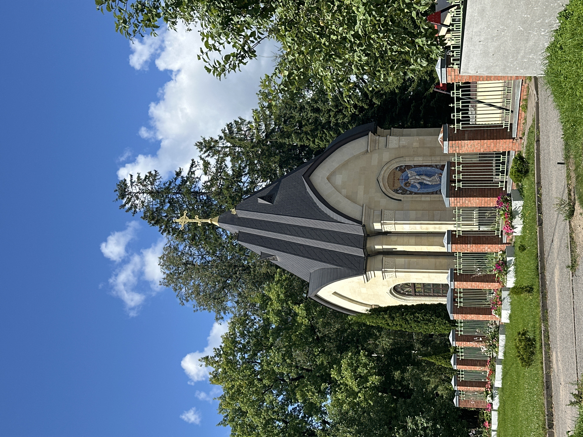 The image shows a small, ornate chapel with a black, multi-faceted roof topped by a golden cross. The chapel is constructed of light-colored stone and features a mosaic on its facade depicting a blue-robed figure with wings. A decorative window with stained glass is visible on the left side of the chapel. The chapel is surrounded by a fence made of brick pillars and light-colored metal railings, with flower beds containing pink and white flowers lining the walkway in front of it. Lush green trees and evergreens form a dense backdrop to the chapel, with a bright blue sky dotted with white clouds visible above. To the right of the chapel, a large tree with green leaves partially obscures the view. This chapel is located at the Vsevolozhsk Cemetery in Vsevolozhsk, Russia.