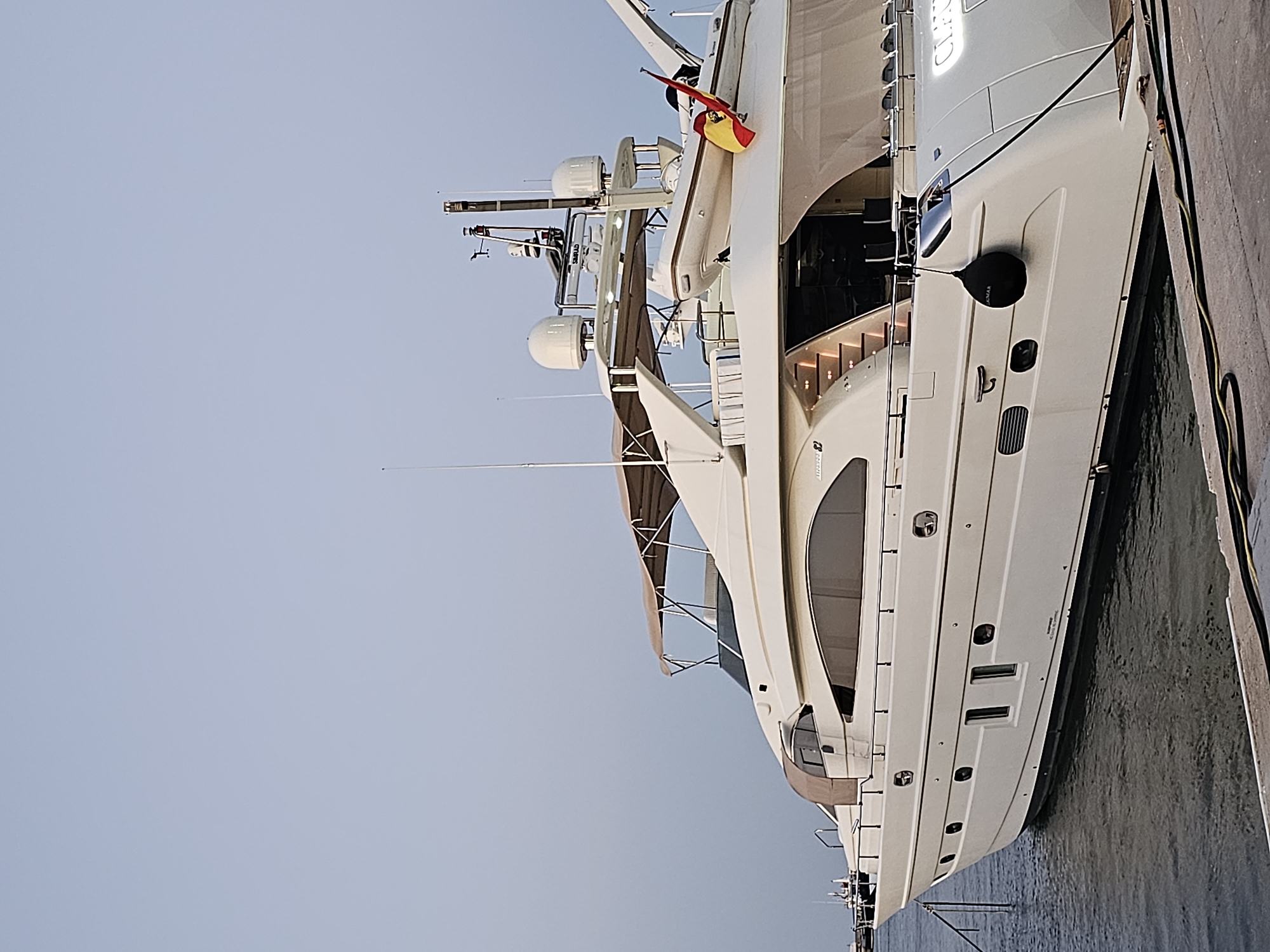 The image shows a large, cream-colored yacht docked at a marina in Dénia, Spain. The yacht is positioned at an angle, with its stern facing the viewer's right. It features multiple levels, large windows, and a covered deck area with seating. A tender boat is visible on the upper deck, and a Spanish flag is flying from a pole at the stern. The yacht is moored to a concrete dock. The water is dark and calm. The sky is a pale, hazy blue, suggesting either early morning or late afternoon light. There are no people visible in the image. The scene appears serene and unoccupied.