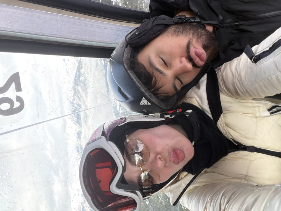 Two individuals are captured in a close-up selfie within a ski gondola cabin in Courchevel, France. The female on the left, wearing a white ski helmet with red/pink accents, ski goggles pushed up, and gold-rimmed light brown sunglasses, pouts her lips towards the camera. She is dressed in a cream-colored padded ski jacket with black stripes and a black neck gaiter. To her right, a male with a dark beard, wearing a black ski helmet with a white logo, has his eyes closed and lips slightly pursed, resting his head close to the female. The scene is viewed through a wet, clear window, showing raindrops or condensation. Outside, snow-covered mountain peaks and an overcast sky are visible. Reflected, inverted text, "es," is faintly discernable on the upper portion of the window.