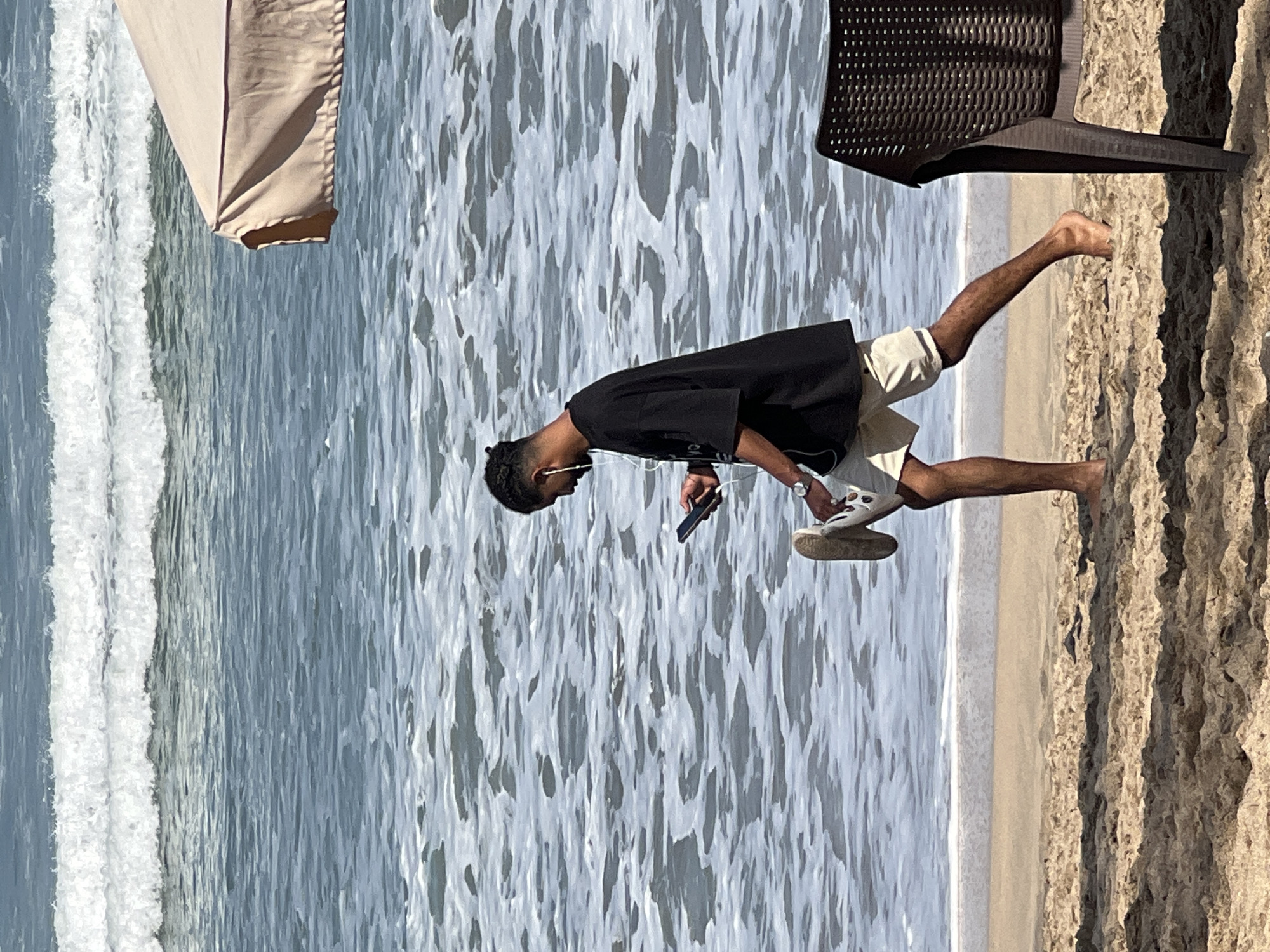 A person walks along the beach in Kuta, Indonesia, holding a smartphone and wearing earphones. The person is dressed in a black t-shirt and light-colored shorts. One shoe is being held in their hand. The background shows the ocean with waves and a beach umbrella. A chair is visible on the right side of the frame. The sand is textured with footprints and indentations. The water is a mix of blue and white from the waves. The lighting suggests it is daytime.