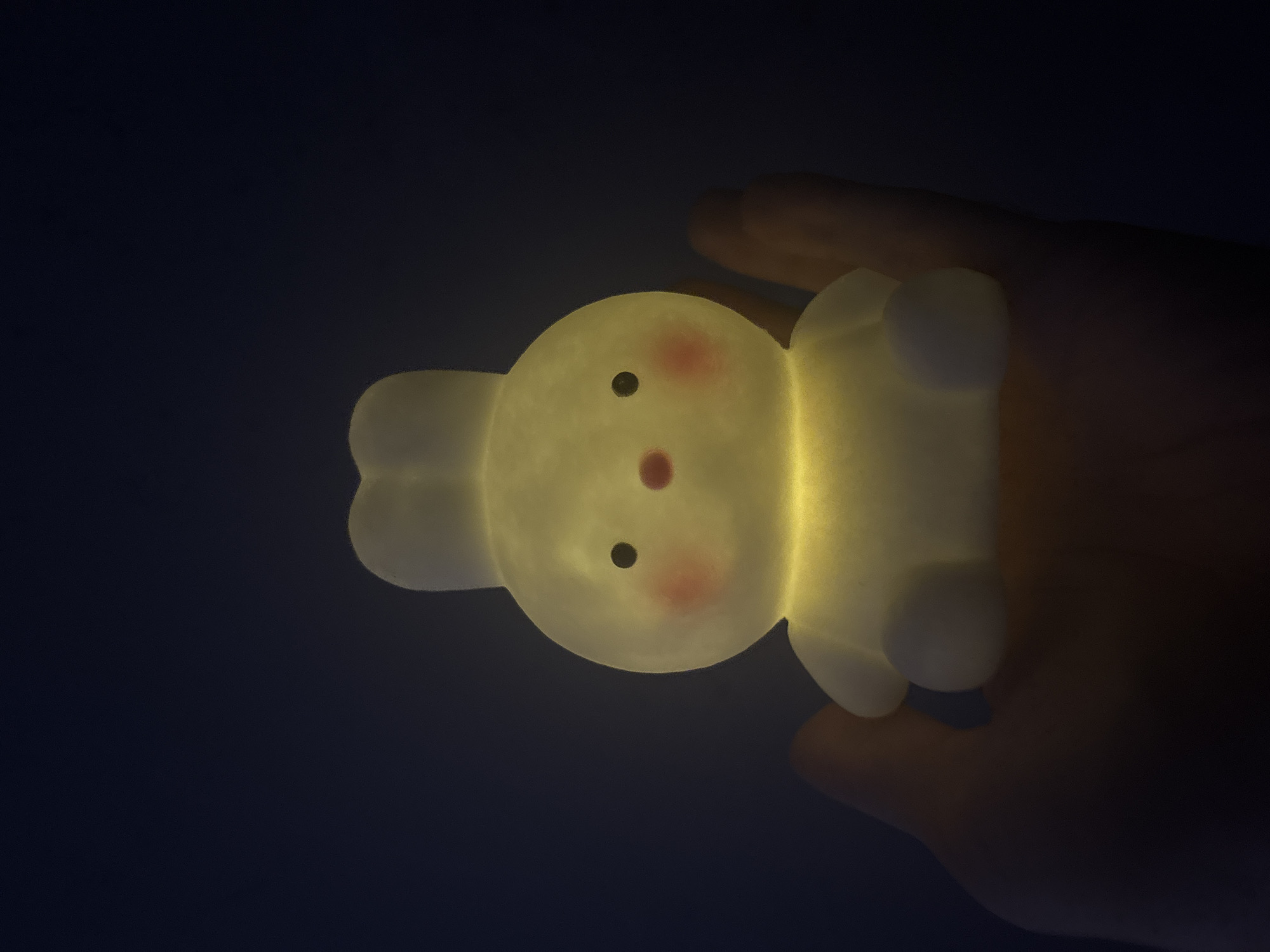 A person's hand holds a small, illuminated bunny-shaped night light. The night light is translucent and emits a warm yellow glow from within. It has two small black eyes and pink blush marks on its cheeks, with a small pink nose. The bunny is sitting upright with its arms and legs tucked in. The background is dark and out of focus, suggesting the image was taken in low light conditions. The location is Unknown, Unknown.