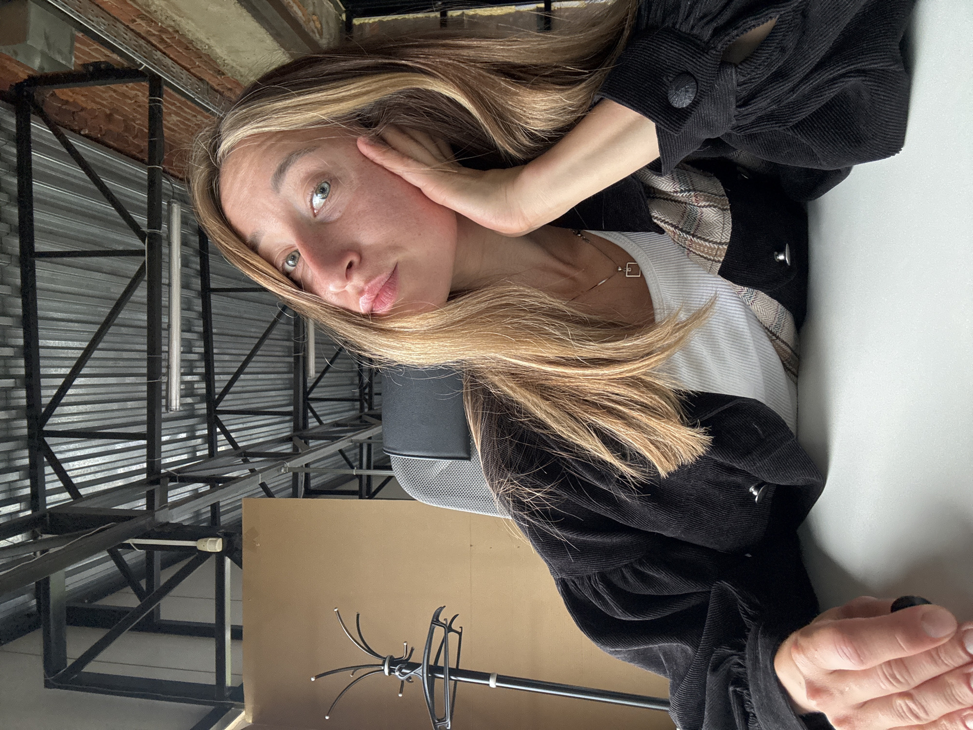 A woman with blonde hair is seated at a white desk in what appears to be an industrial-style space. She is resting her right cheek on her right hand and looking towards the camera with her mouth slightly open. She is wearing a black corduroy jacket over a white tank top with a plaid detail. In the background, a coat rack is visible to the left, and the ceiling features exposed metal beams and corrugated metal roofing. The environment suggests a renovated industrial building, possibly an office or studio space, located in the city of Chaikovsky, Russia.