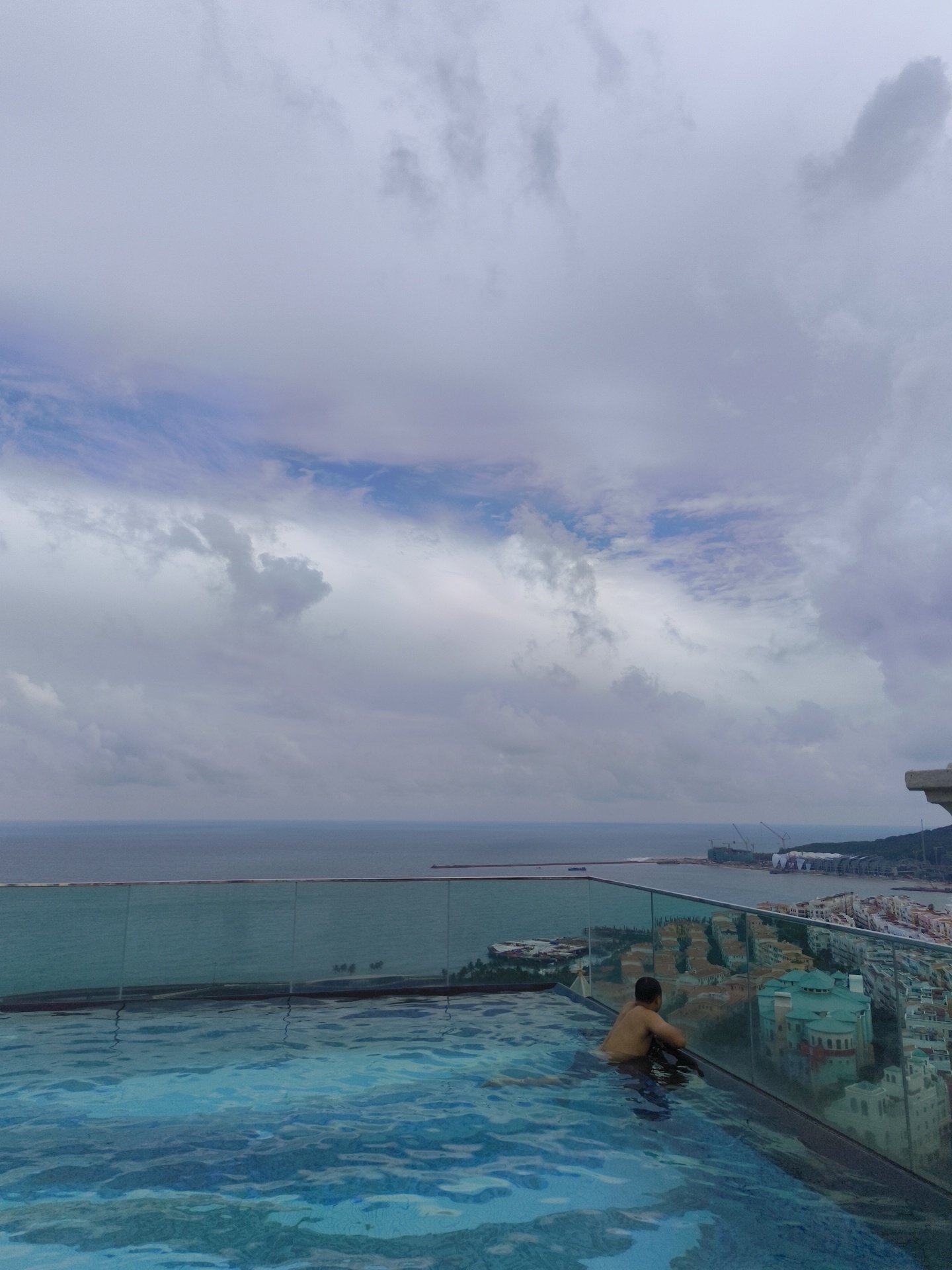 A person is partially submerged in a rooftop infinity pool in the city of Phu Quoc, Vietnam. The pool water is blue and rippled. The person is facing away from the camera, looking out at the ocean. The ocean is calm and gray, with a cloudy sky above. In the background, there is a city with many buildings, some of which are colorful. There are also cranes and construction activity in the distance. The overall mood of the image is peaceful and serene.