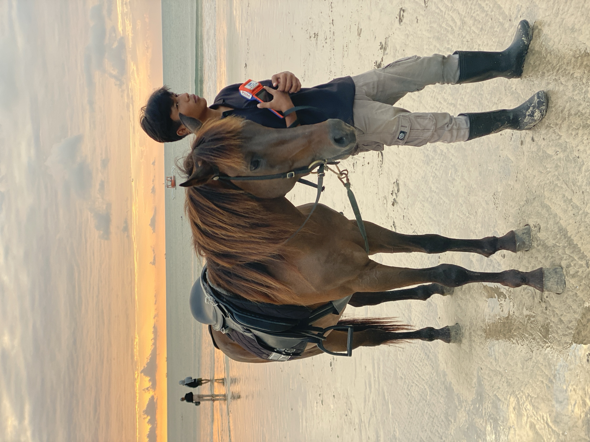 A young man stands beside a brown horse on a wet, sandy beach in Kuta Selatan, Indonesia, during sunset. The man, wearing a dark t-shirt, cargo pants, and black rubber boots, holds a small orange and red device in his right hand. The horse is saddled with a black saddle and pad. In the background, the ocean stretches out to the horizon under a sky filled with colorful clouds painted with hues of orange, yellow, and pink. Two figures, a man and a woman, are walking in the shallow water further down the beach, and a small boat is visible in the distance. The wet sand reflects the colors of the sunset.