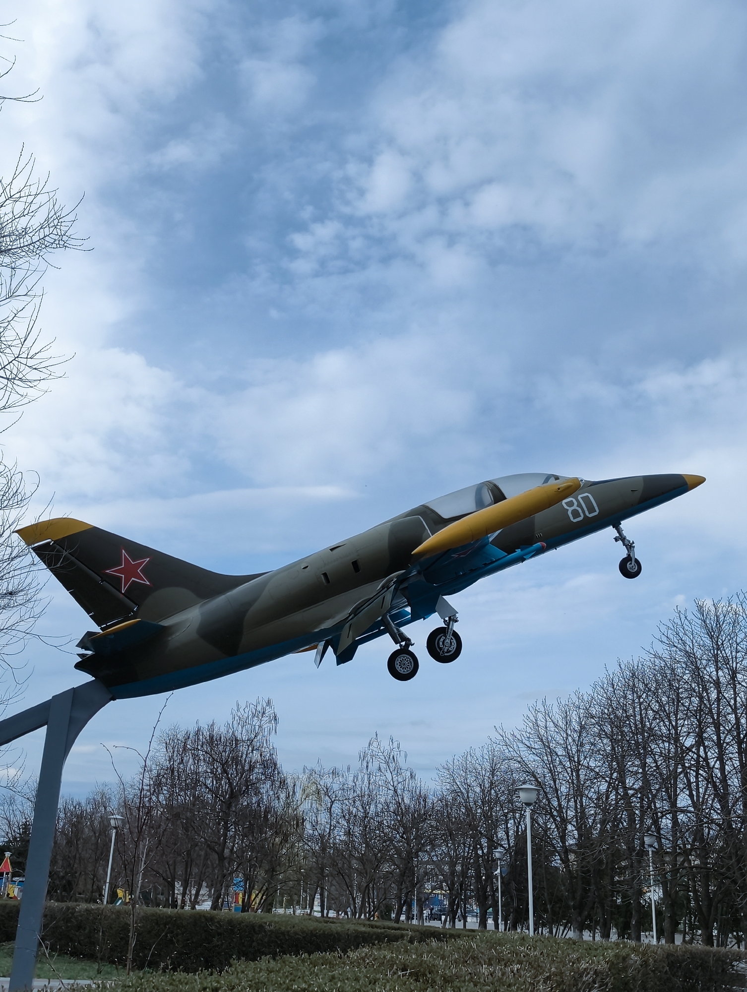 The image shows a military aircraft, specifically an L-39 Albatros trainer jet, mounted on a metal support stand as a monument in a park. The plane is painted in camouflage colors with a red star on the tail and the number "80" on the nose. The landing gear is deployed. The background consists of a cloudy sky and bare trees indicating a season like late autumn or early spring. The monument is located in a park setting in the city of Balashov, Russia. In the park, there are other trees, bushes, park lighting fixtures, and hints of a playground in the distance.