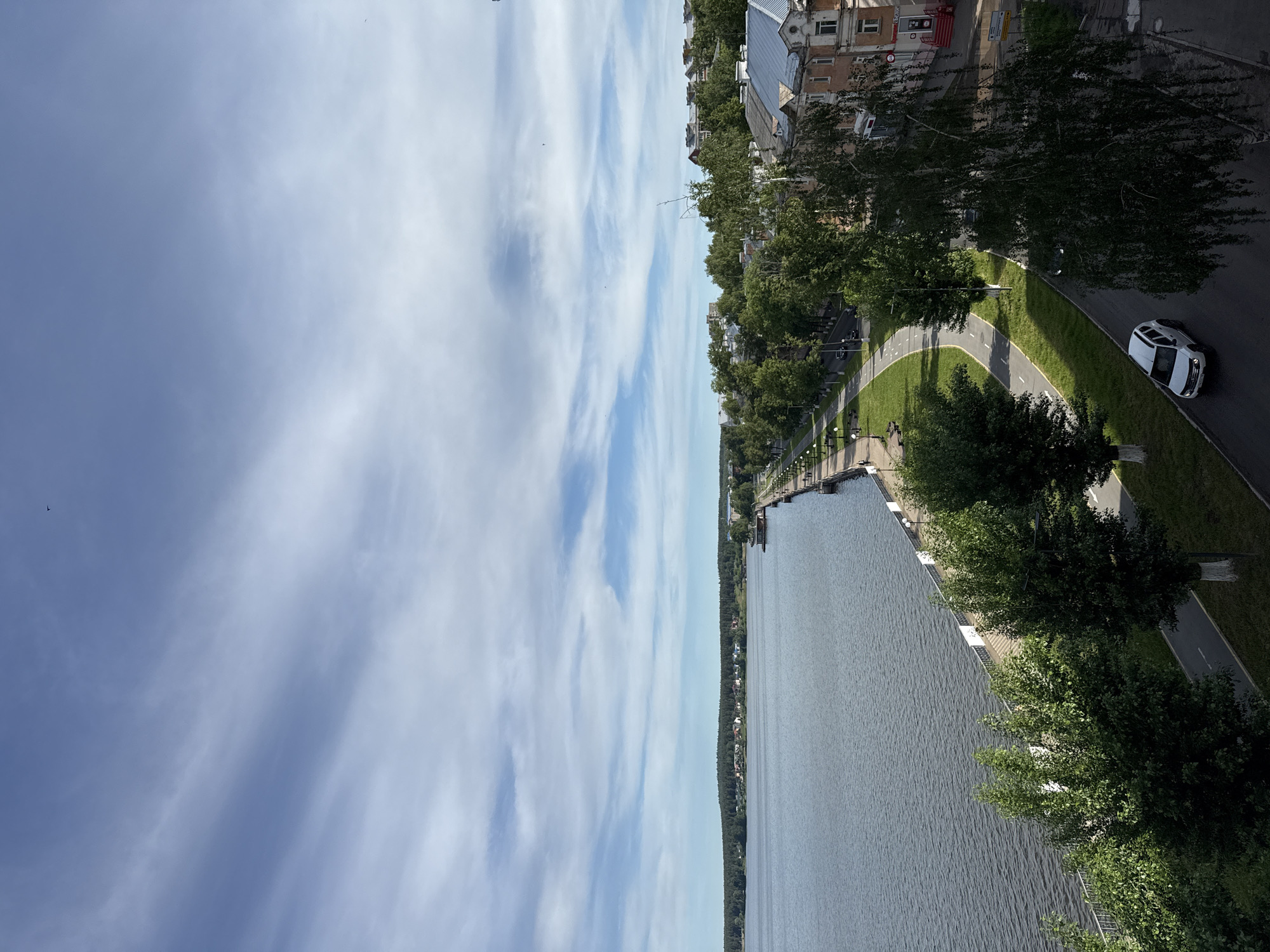 The image shows a view overlooking a riverfront area in Votkinsk, Russia. The sky is light blue with streaks of clouds. The river, a dark blue-gray color, fills the right half of the frame. A paved walkway runs alongside the river, separated by a line of trees. A grassy area runs alongside the walkway, with a road further in. A single white car is on the road. Buildings, possibly residential, are visible further back. A few individuals can be seen walking along the riverfront walkway. Streetlights are placed along the walkway.