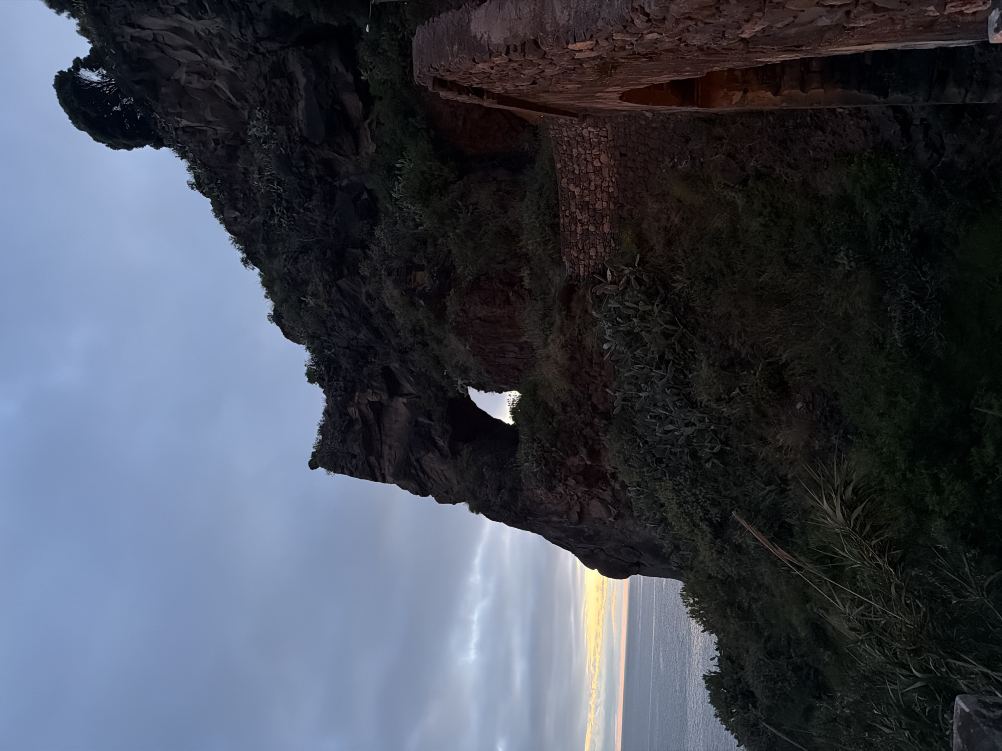 The image shows a rock formation with a hole in it, set against a cloudy sky. The location is Ponta do Sol, Portugal, based on the landscape. A stone structure is built along the top edge of the cliff. The rock formation is covered in green vegetation on its edges. The ocean is visible at the bottom of the image.