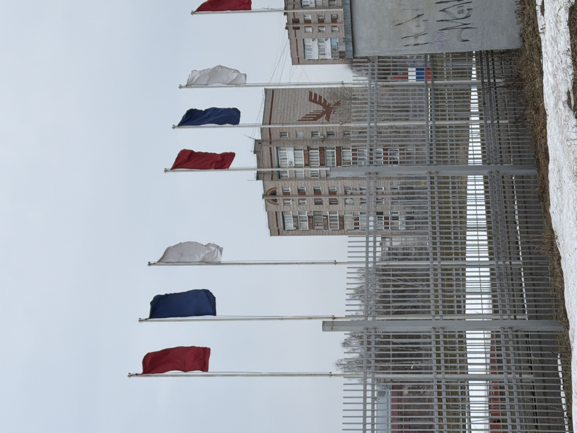 The image depicts the Monument "Anchor" in Votkinsk, Russia. Seven flags in the colors of the Russian flag (red, white, and blue) are visible on flagpoles in the foreground. In the background, a building with a Soviet-era emblem is visible, partially obscured by a metal grid fence. A concrete structure with graffiti is visible in the upper right corner, and snow covers the ground. The sky is overcast.