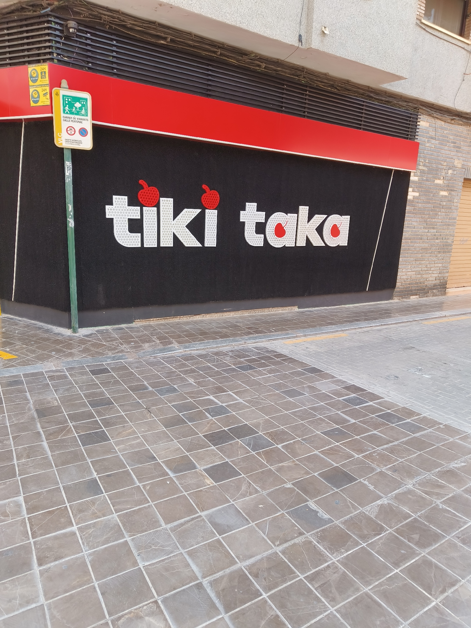 The image shows the exterior of a building with a black facade and large white lettering that reads "tiki taka." Two red apple graphics are positioned above the letters. A red horizontal band runs along the top of the facade. To the left, a yellow sign with green lettering and graphics indicating a pedestrian street is attached to a green pole. The building appears to be located on a street with a tiled sidewalk. The overall scene is a street view of a commercial establishment in the Benimàmet-beniferri district of Valencia, Spain.