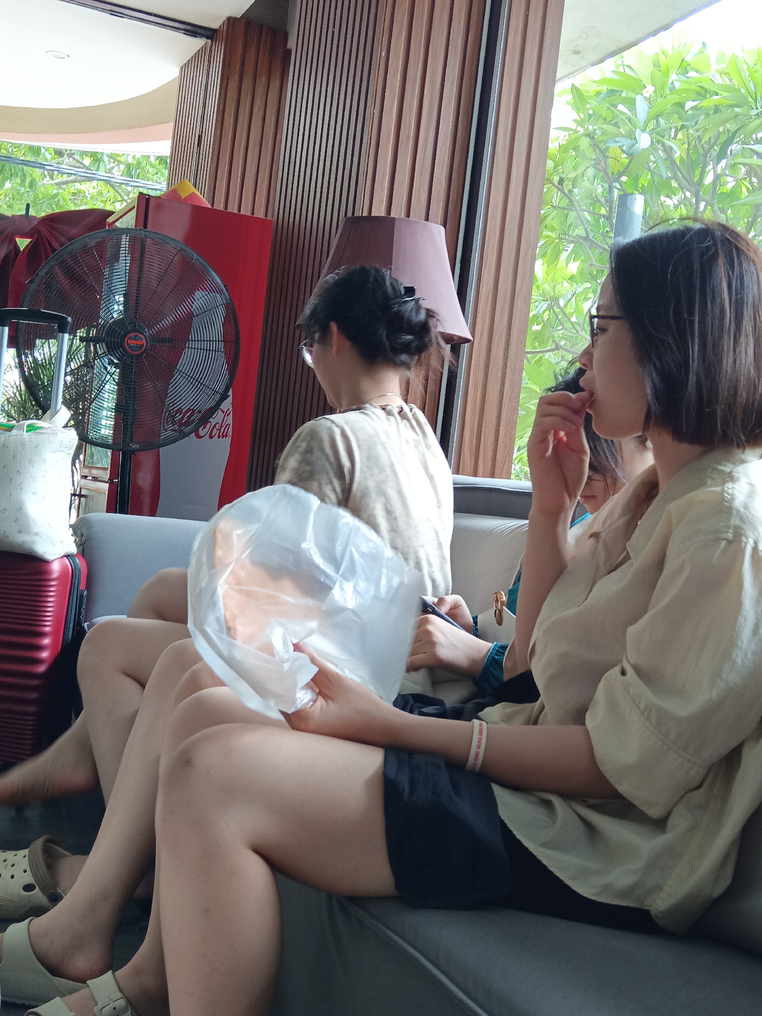 The image depicts three individuals, two women and one partially visible person, seated on a sofa. The woman in the foreground, on the right, has short dark hair, wears glasses, and is seen eating. She is dressed in a light-colored collared shirt and dark shorts. To her left, another woman with dark hair tied back is seated, facing away from the camera. She is wearing a light-colored long-sleeved shirt. A third person is partially visible behind them, wearing a teal garment.
The environment appears to be an indoor waiting area or lounge. Behind the sofa, a tall red cabinet with a Coca-Cola advertisement is visible, alongside a large black oscillating fan. Wooden paneling adorns the wall. Large windows offer a view of lush green trees and foliage, suggesting a tropical setting. Two suitcases, one red and one a lighter color, are positioned to the left of the sofa, indicating travel. The overall atmosphere is casual and relaxed. This scene could be located in Đà Nẵng, Vietnam, a coastal city known for its resorts and tropical climate, which would align with the observed environment.